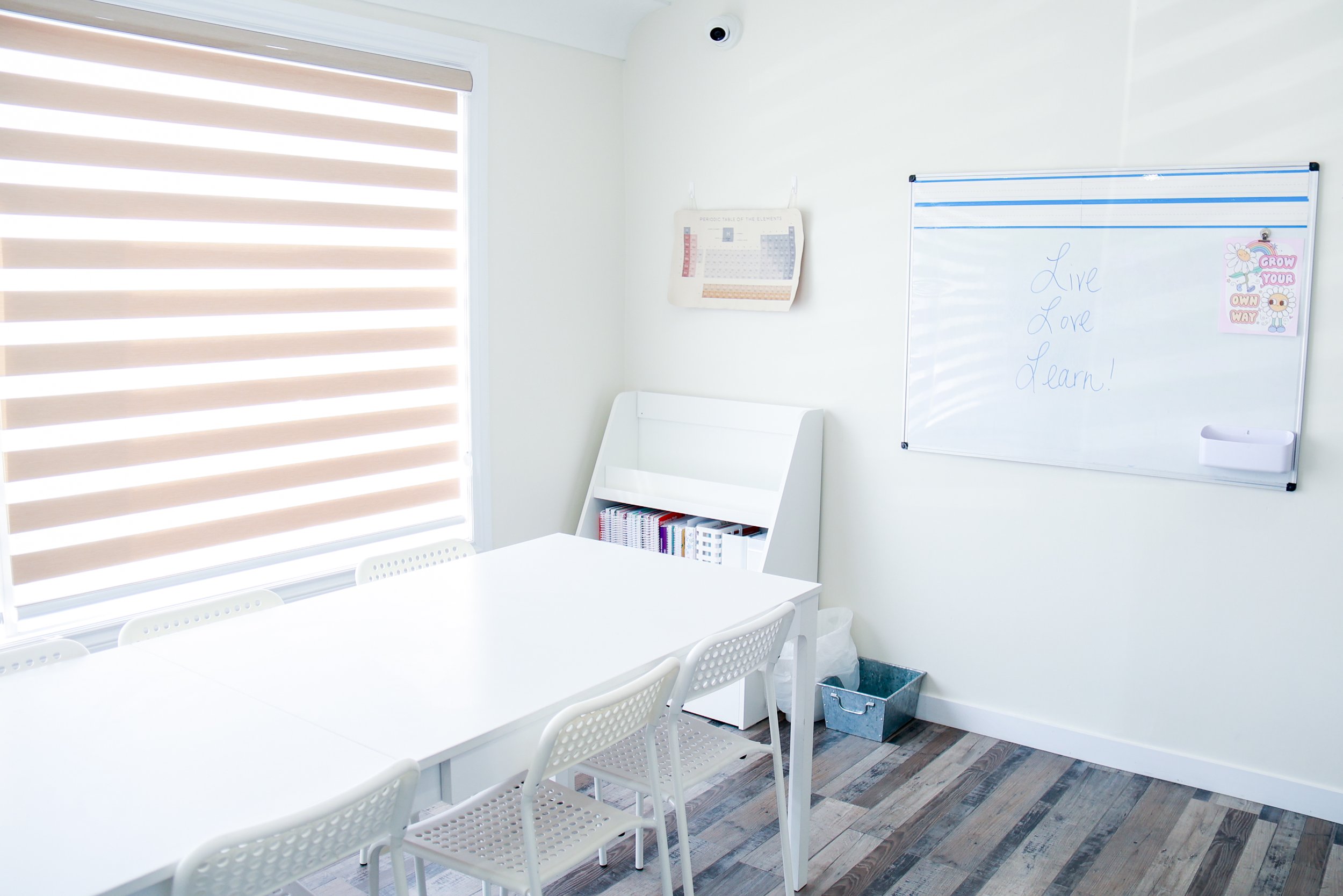 Empty classroom with white tables and chairs, whiteboard with the words "Live Love Learn!", window with beige blinds, bookshelf, and educational posters on white walls.