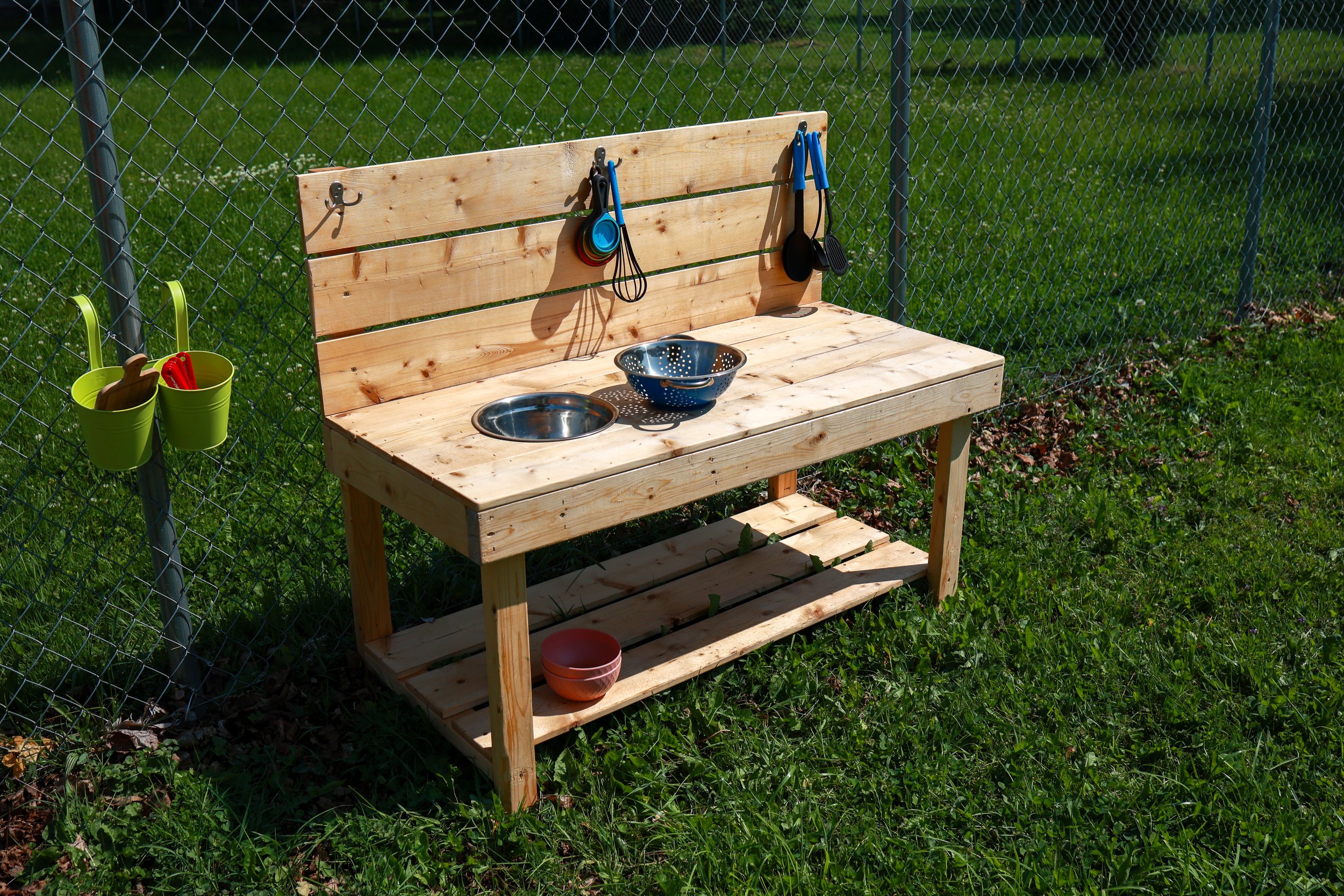 Wooden outdoor play kitchen with utensils and bowls, attached to a chain-link fence in a grassy yard.