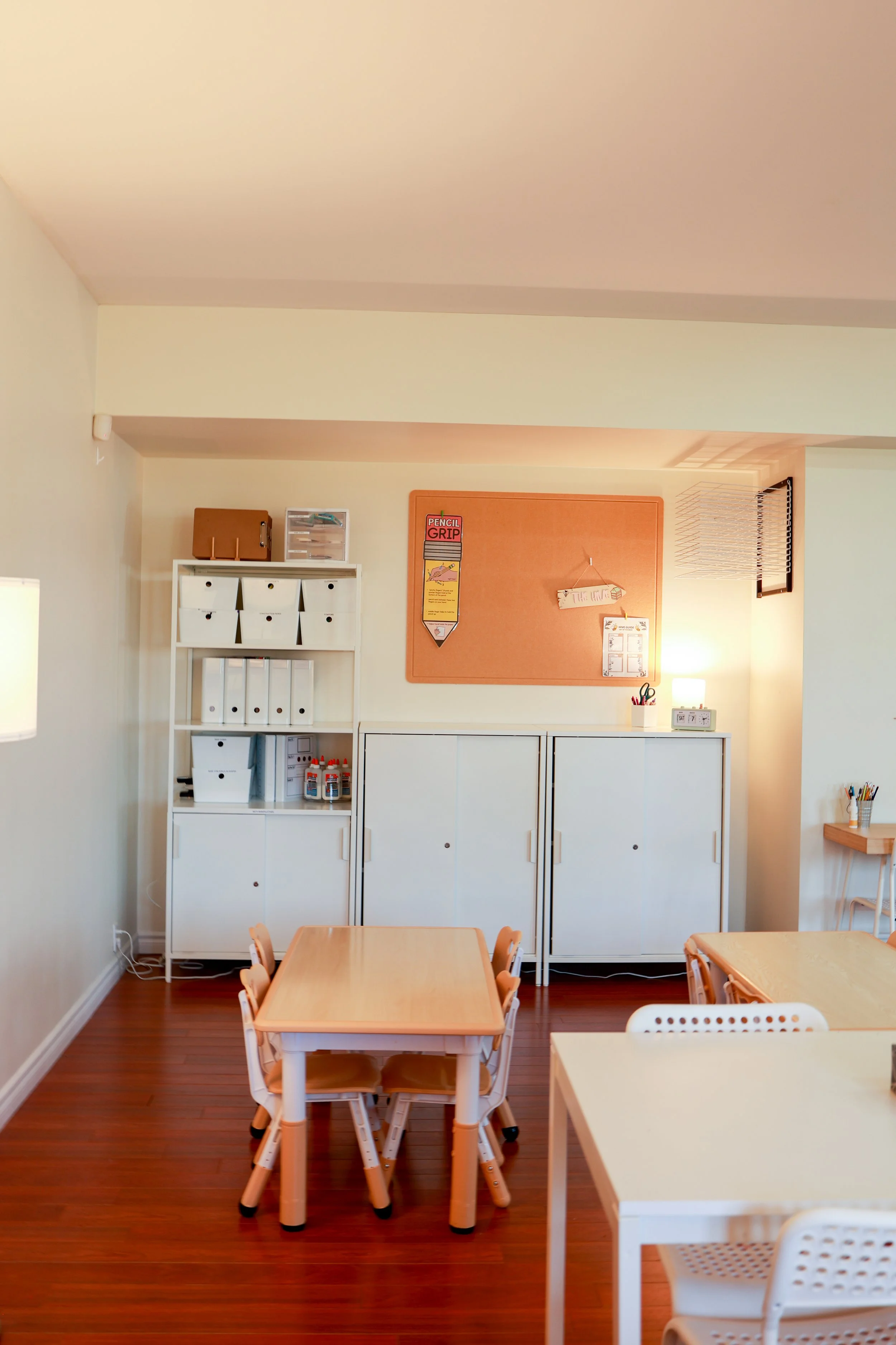 A children's classroom corner with small tables and chairs, white storage cabinets, and a corkboard on the wall.