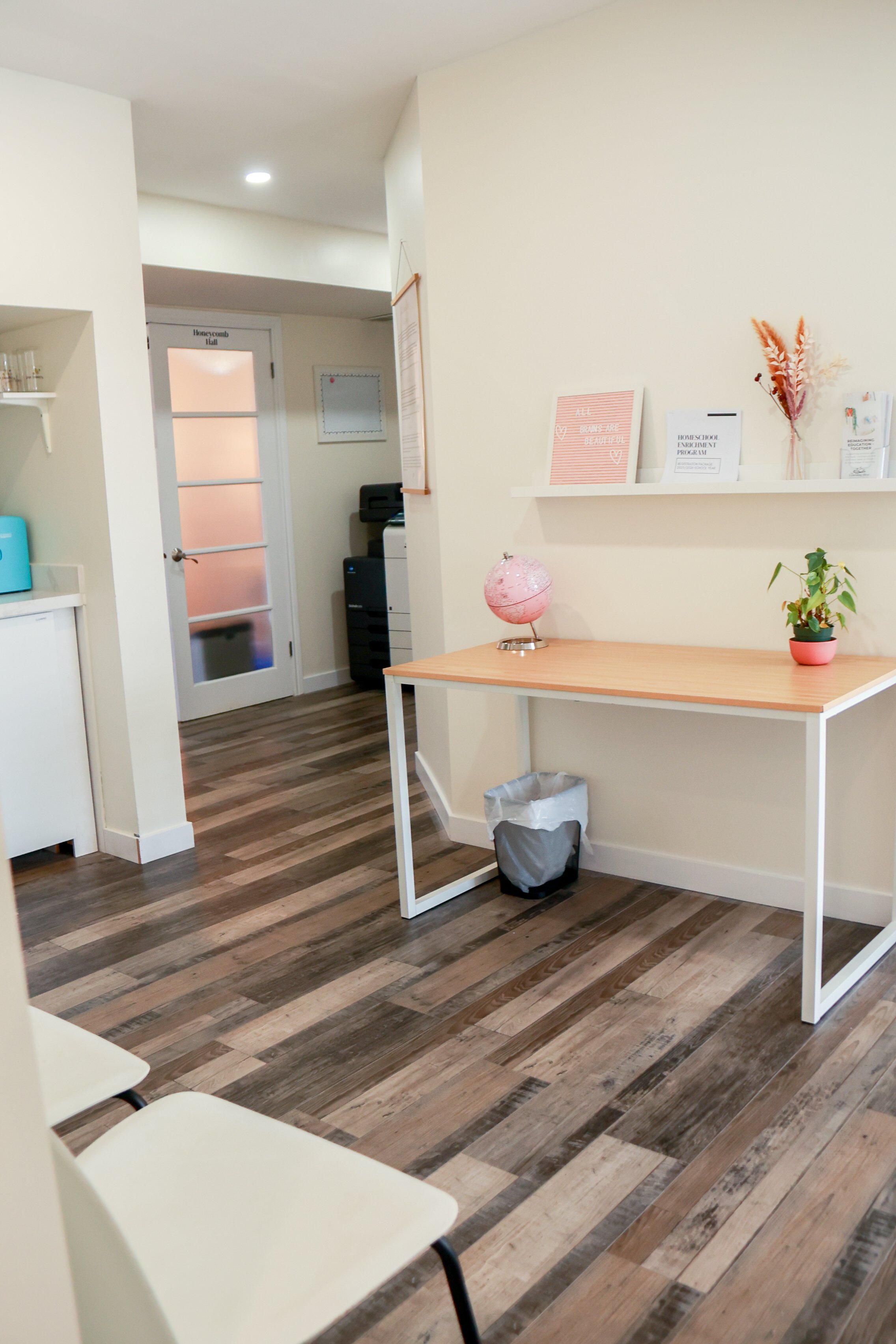 Interior space with a wooden table, small pink globe, potted plant, and decorative items on a wall shelf. Background features a frosted glass door, a corner with a printer, and seating with cream-colored chairs.