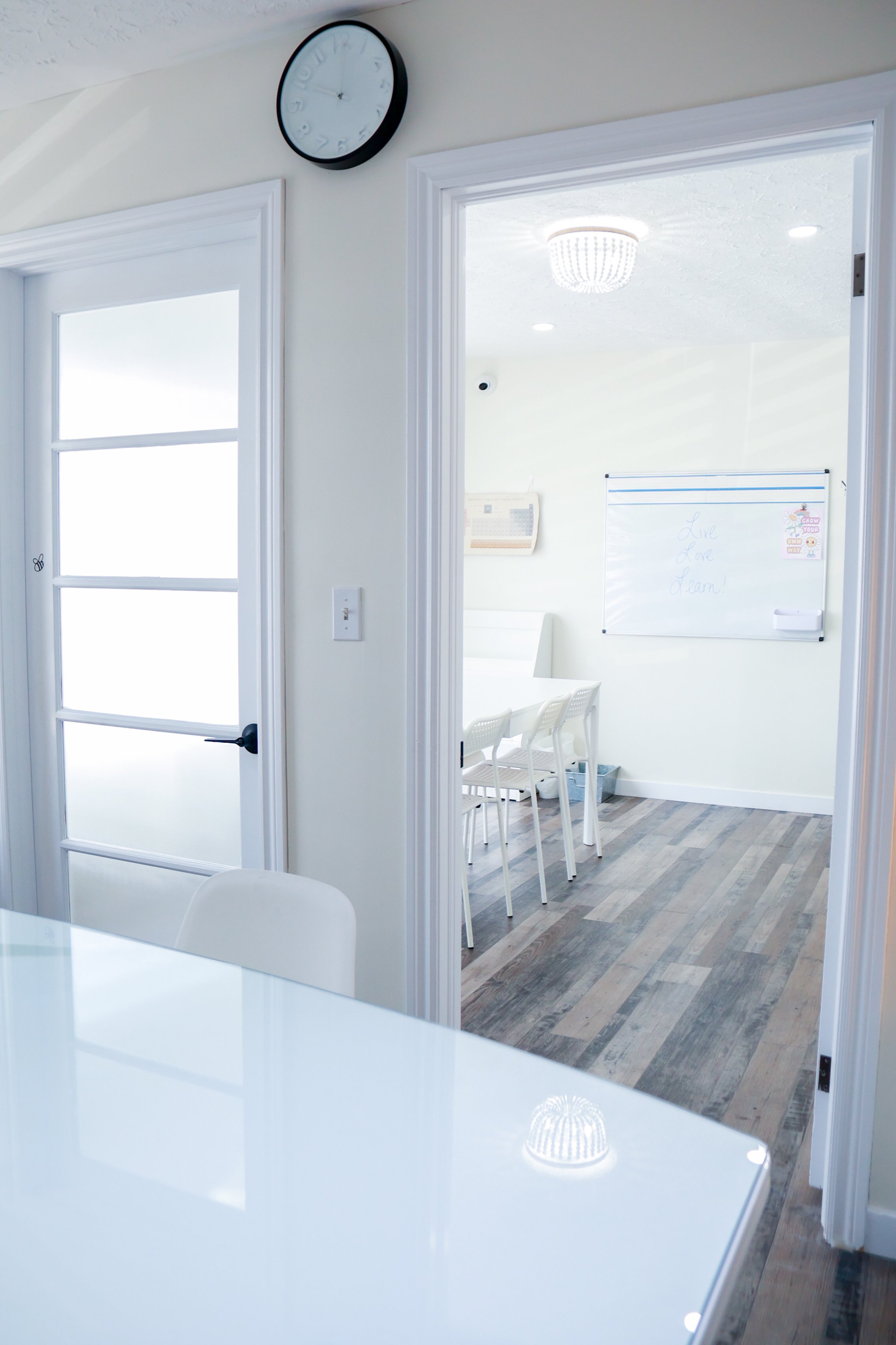 A view through an open doorway into a bright room with white walls, featuring a whiteboard with writing and chairs around a table, with a wooden floor.