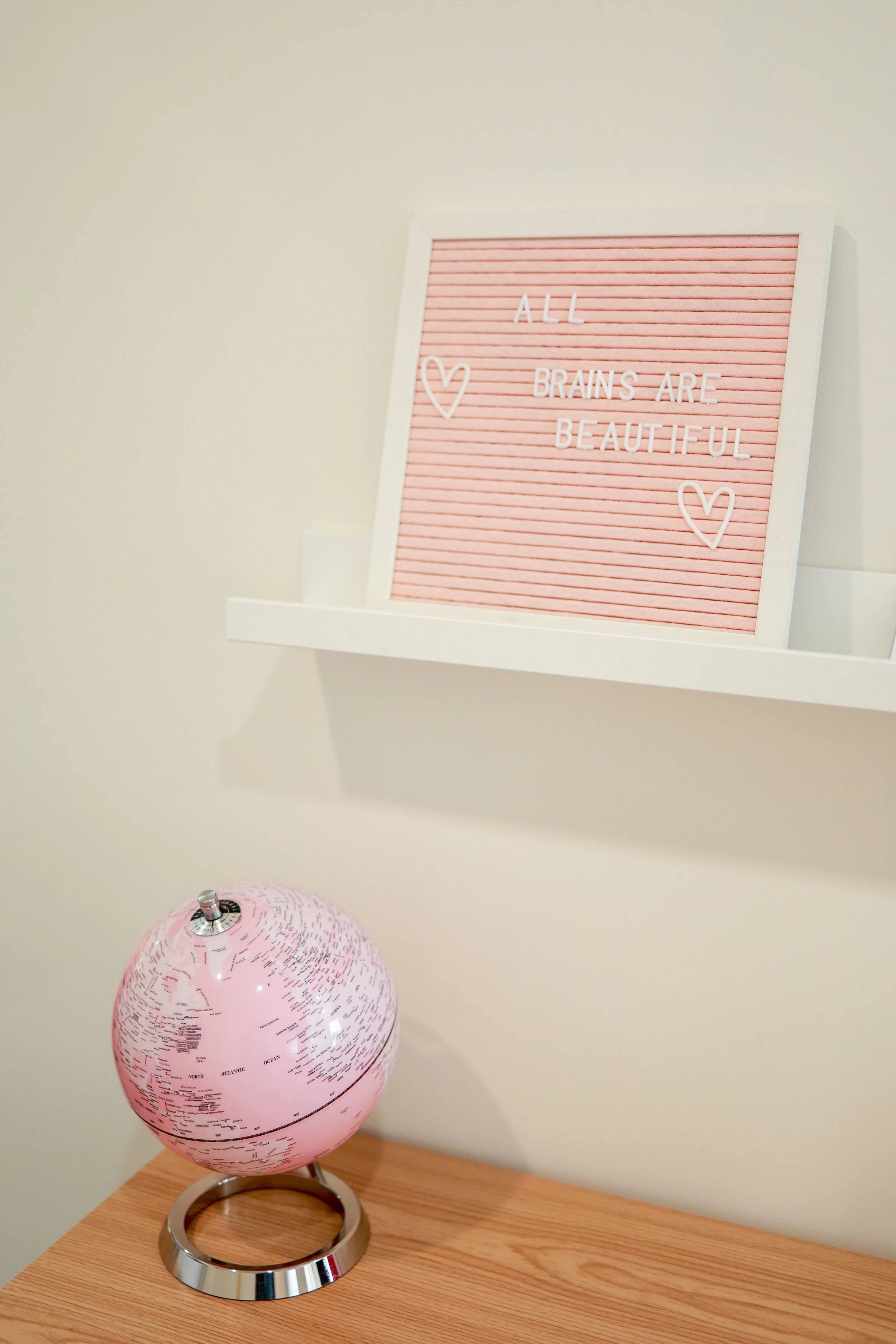 A pink globe and a pink letter board on a white shelf against a beige wall, with a pink quote sign reading "All brains are beautiful" and hearts.