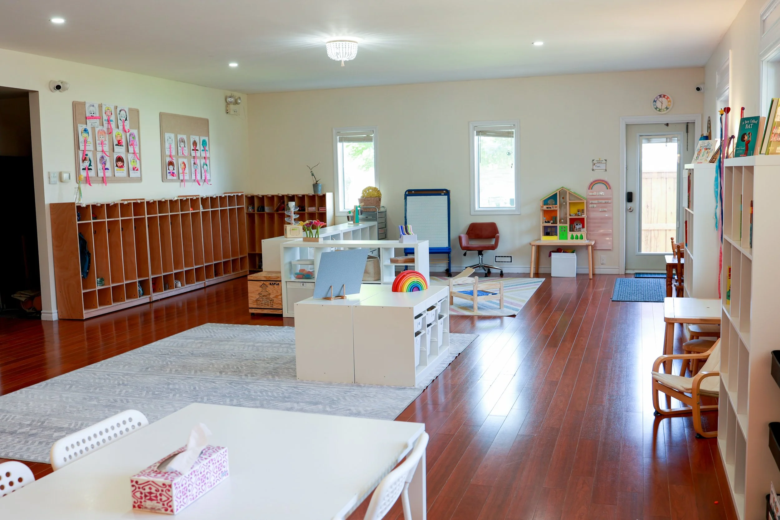 Empty classroom or daycare room with wooden flooring, white walls, and children's furniture and toys. There are shelves, storage cubbies, colorful artwork on the walls, and windows letting in natural light.
