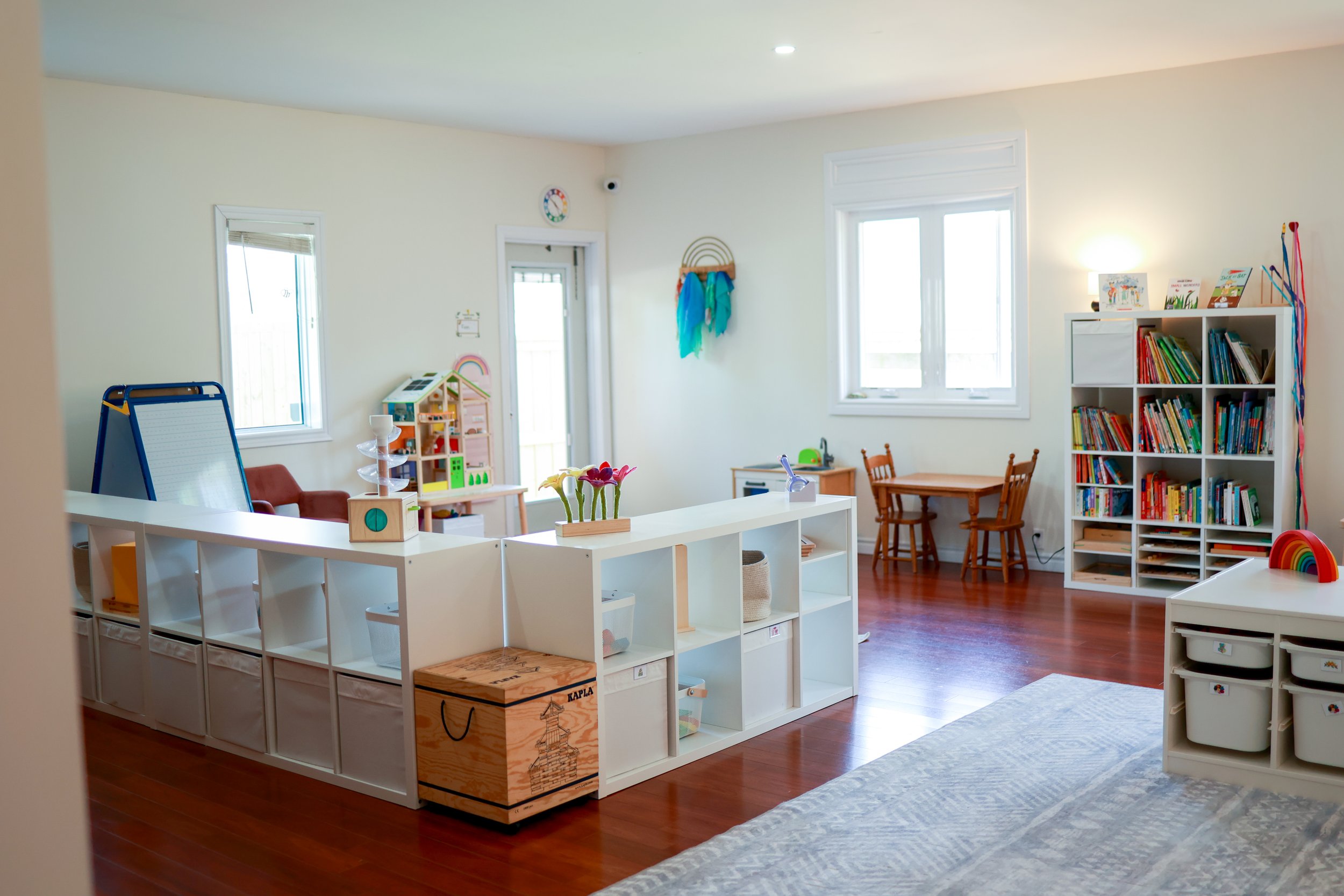 Colorful children's playroom with windows, bookshelf filled with books, small table with chairs, and various toys and art supplies.