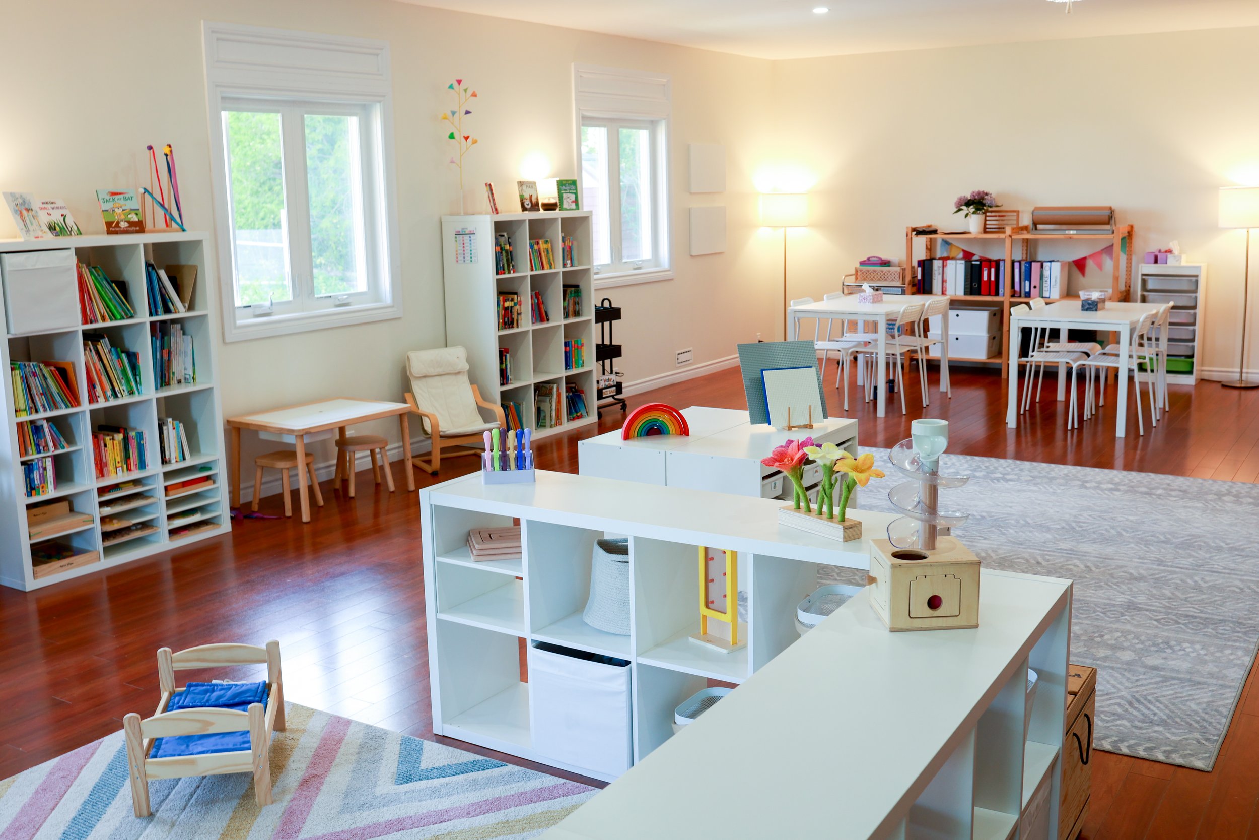 A colorful classroom with wooden floors, white bookshelves filled with books, small tables and chairs, and decorative items like flowers, rainbow, and art supplies.