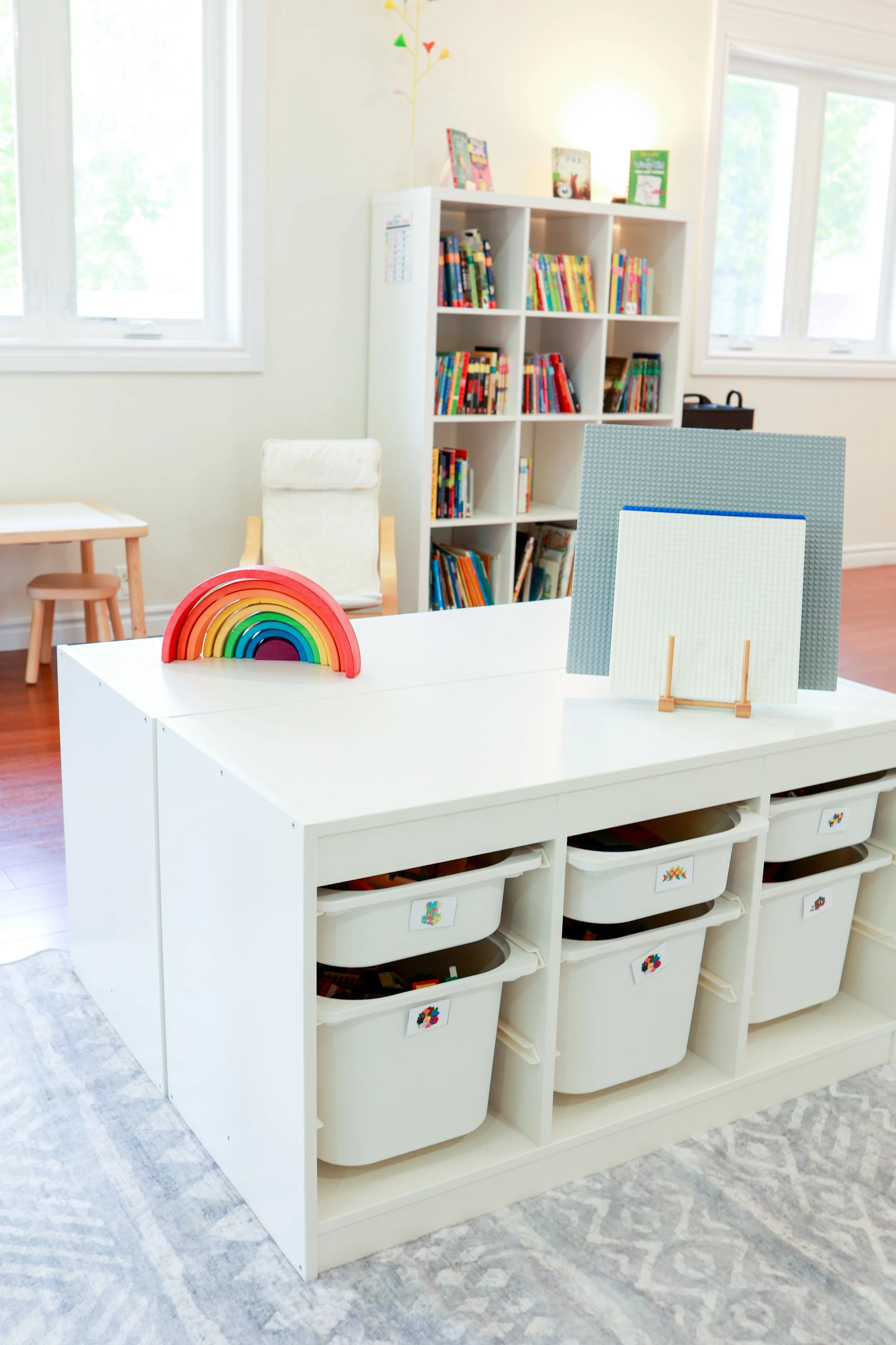 Child's playroom with a white storage unit holding bins, a rainbow stacking toy, and a bookshelf filled with colorful books.