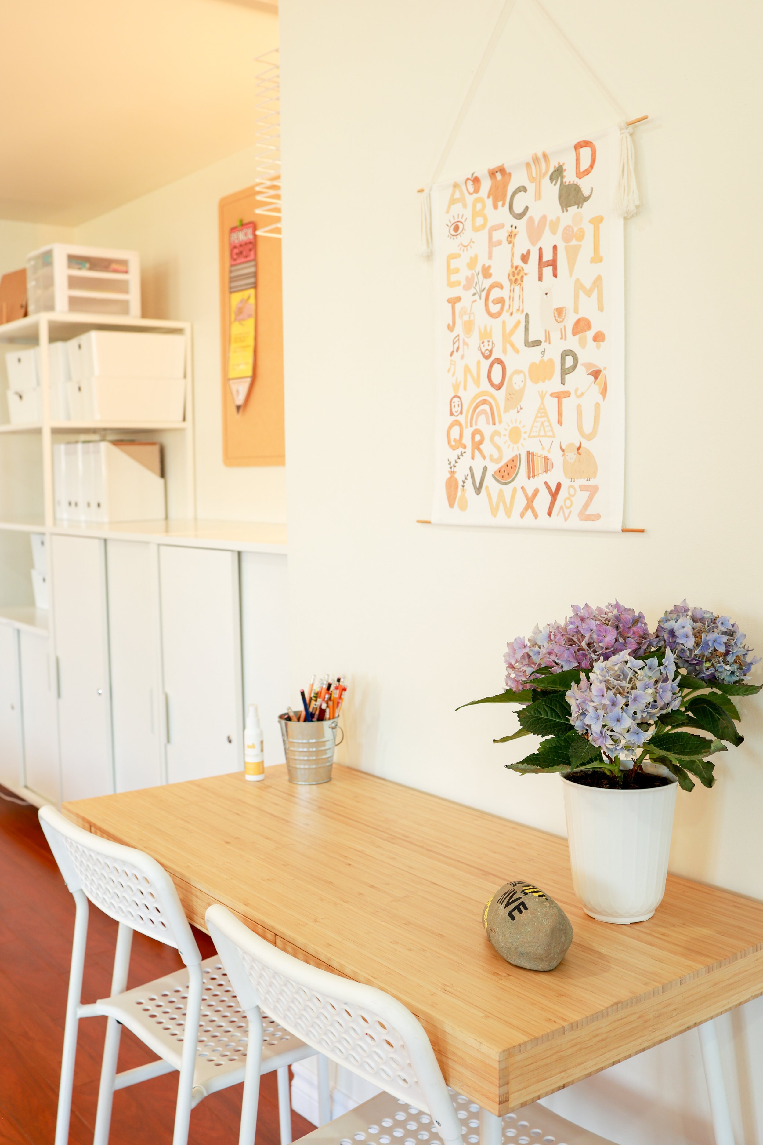 A bright, cozy room with a light wood table, two white chairs, a potted purple-red hydrangea plant, and a wall hanging of the alphabet with illustrations. There are storage cabinets and a bulletin board in the background.