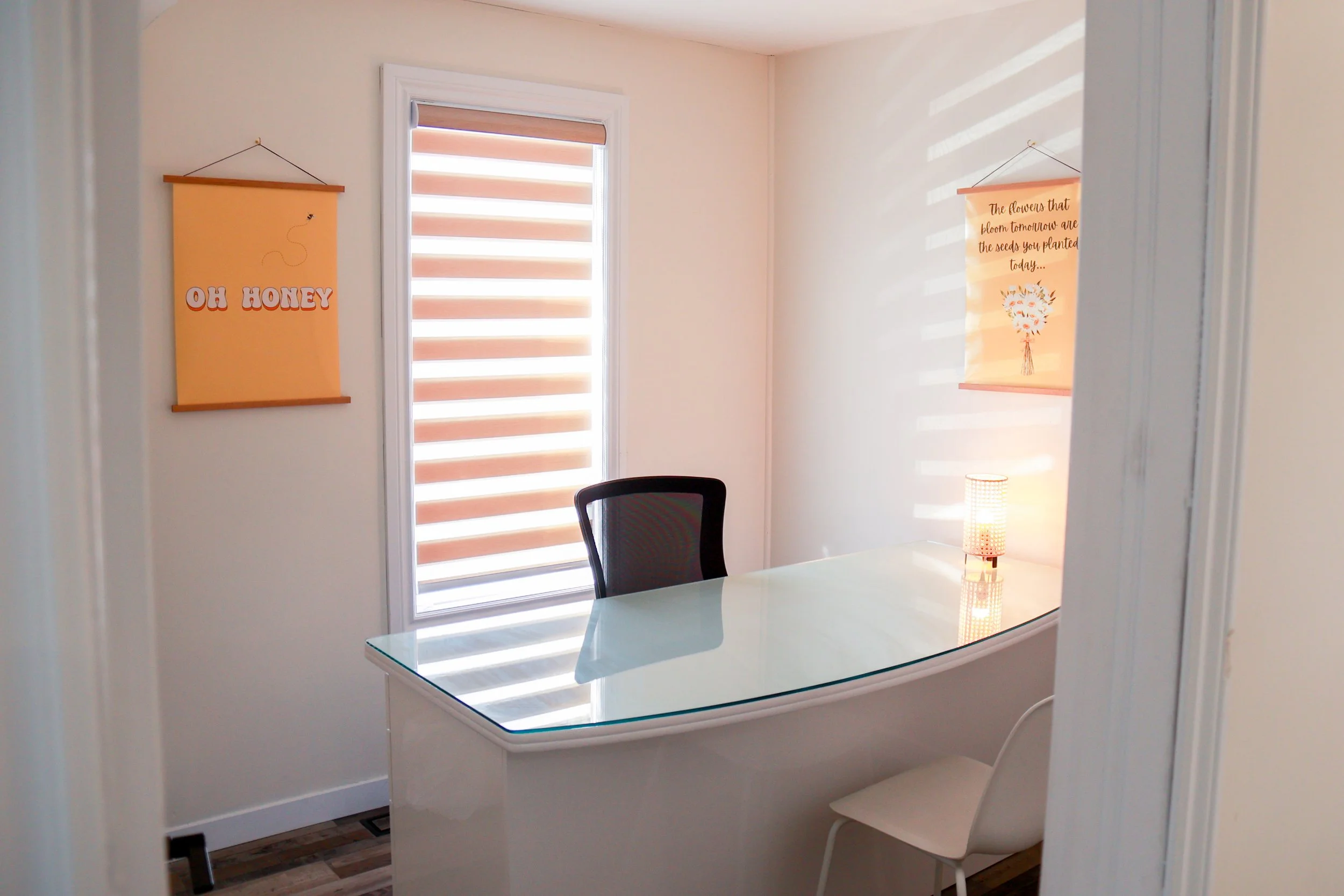 A small office with a glass desk, a black office chair, a white chair, a window with striped blinds, two hanging wall art pieces, and a small table lamp.