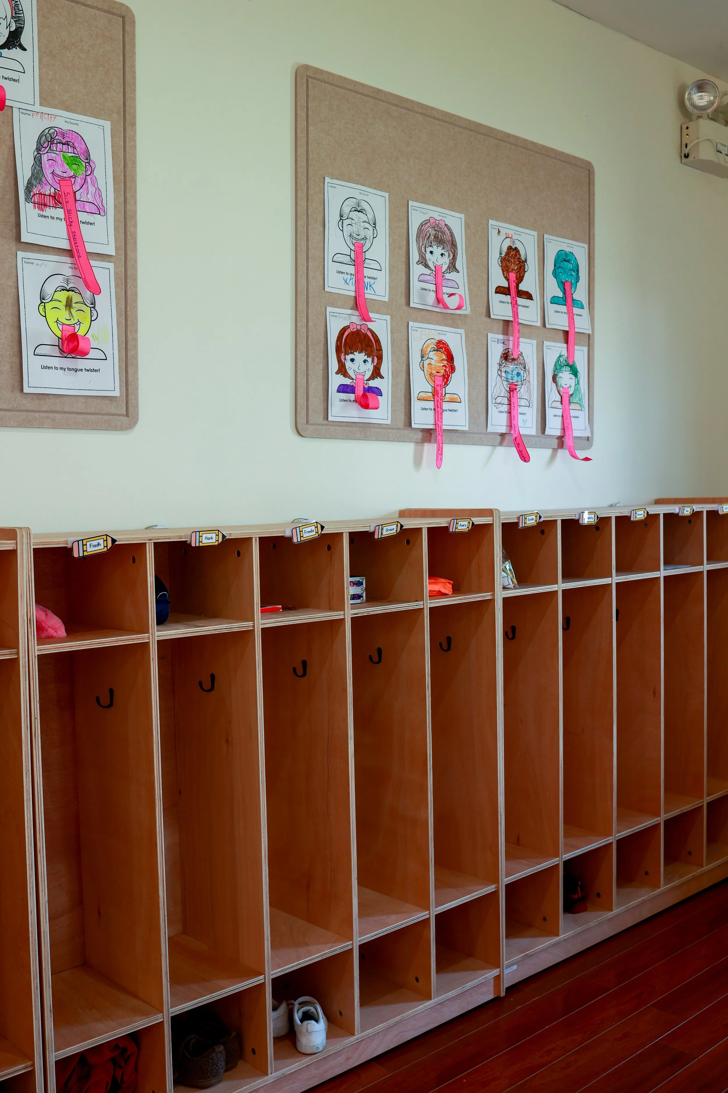 Empty wooden cubbies in a classroom with colorful hooks and labels. Above, there are two bulletin boards displaying illustrated faces with pink tongue-and-mouth cutouts used for a speech therapy or language activity.