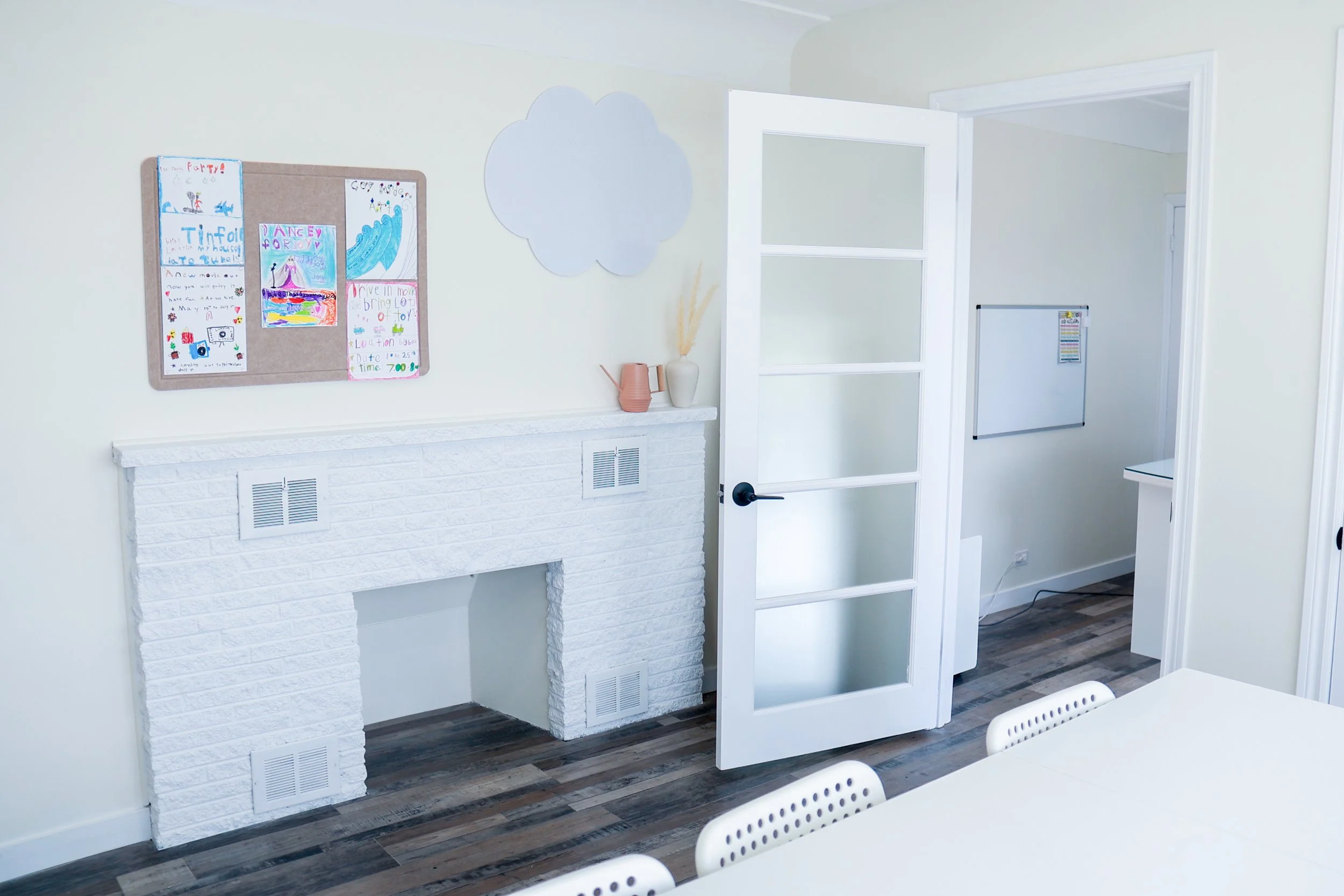 A bright, modern room with baby blue walls, a white brick fireplace, and a glass-paned door. There are children's drawings on a bulletin board, a decorative cloud-shaped wall piece, and a whiteboard in the background.