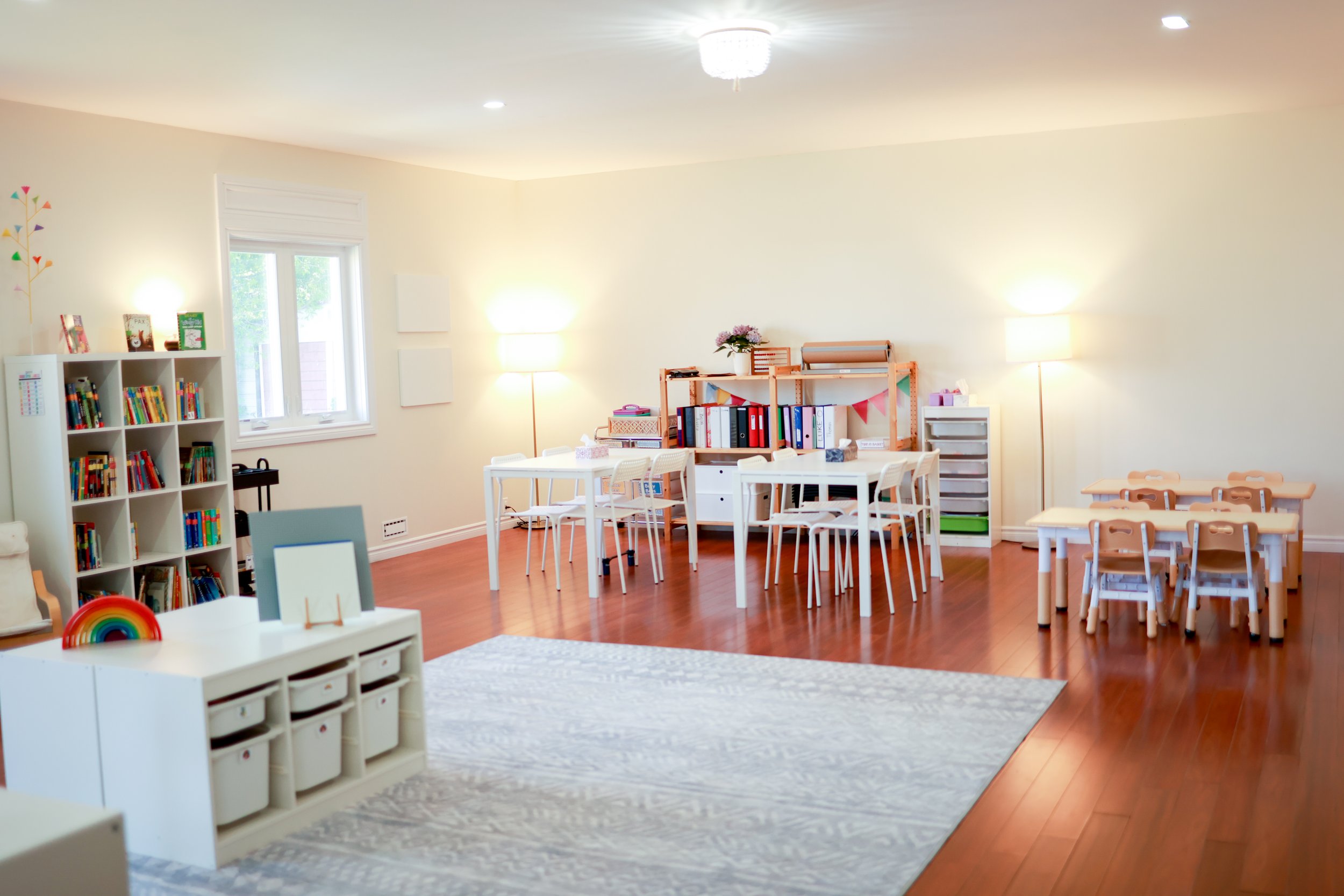 Bright children's classroom with small tables and chairs, a bookshelf with books, storage bins, lamps, and colorful decorations.