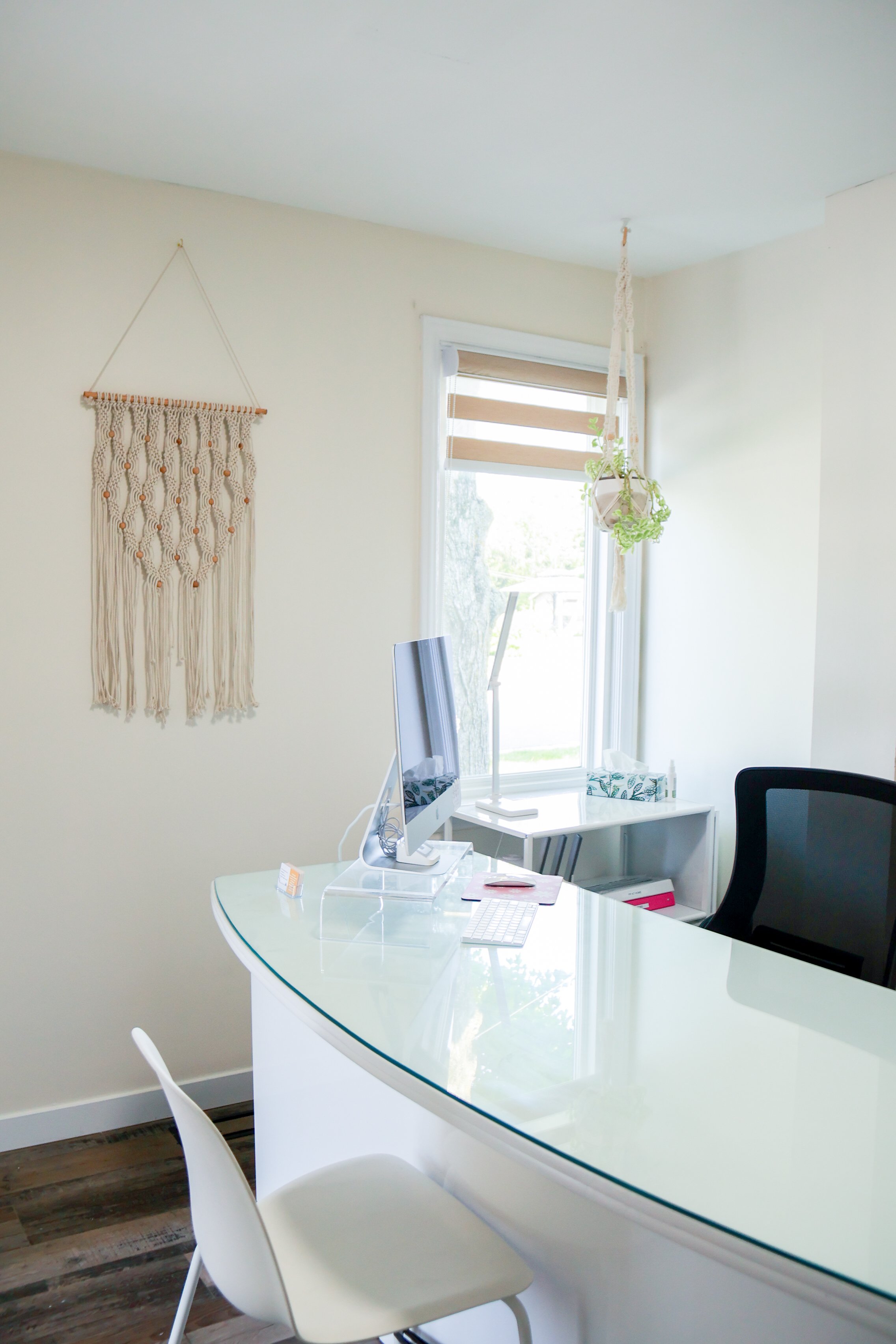 Home office with a white desk, black office chair, and a computer, near a window with natural light and hanging plants, decorated with a wall tapestry.