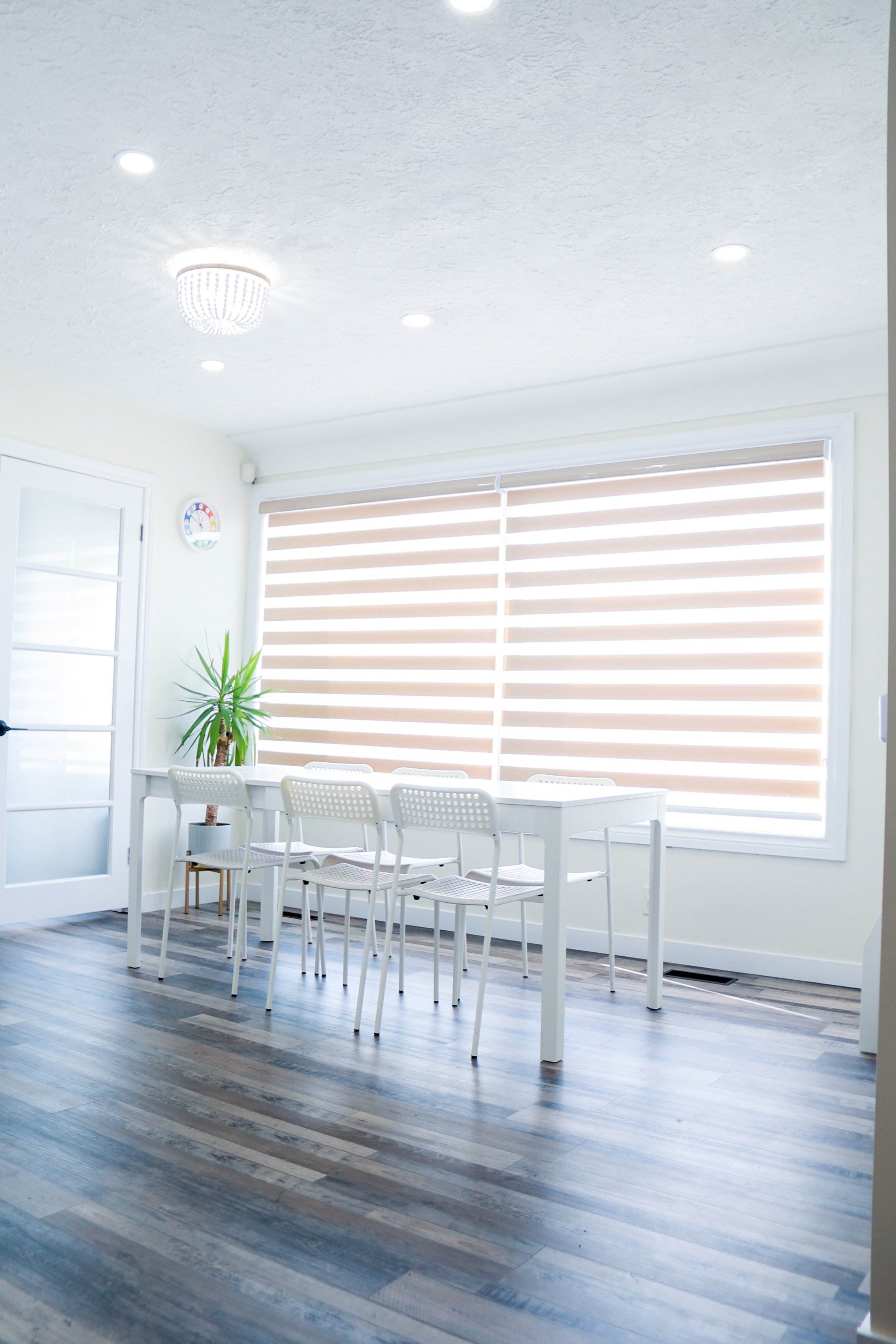 Bright dining room with white table and six white chairs, large window with beige horizontal blinds, potted plant on the table, wooden floor, and white walls.