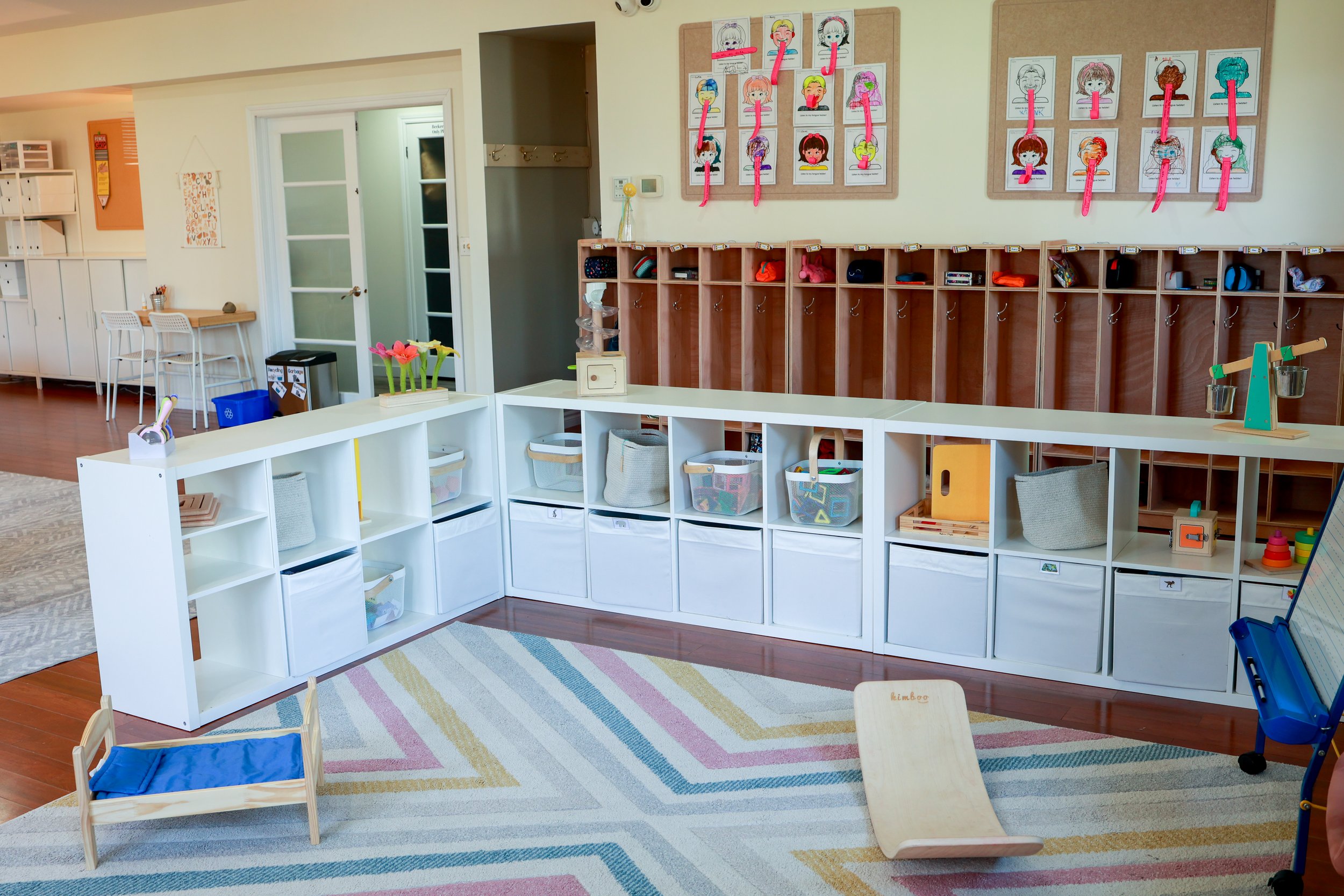 Classroom with cubbies, artwork, and children's toys, arranged on a colorful rug.