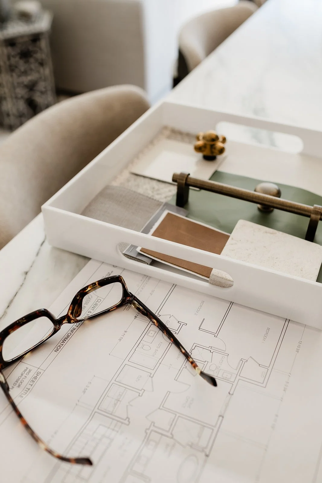 Architectural blueprints placed on a white table with a pair of tortoise shell eyeglasses on top, alongside a white box containing various material samples and design elements for a home renovation or construction project.