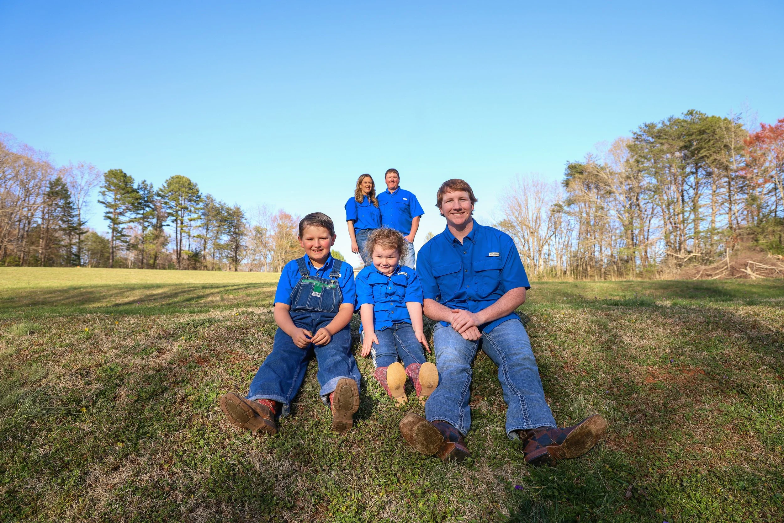 Family of five sitting on grassy hill in front of trees and blue sky, all wearing blue shirts and jeans, smiling at camera.