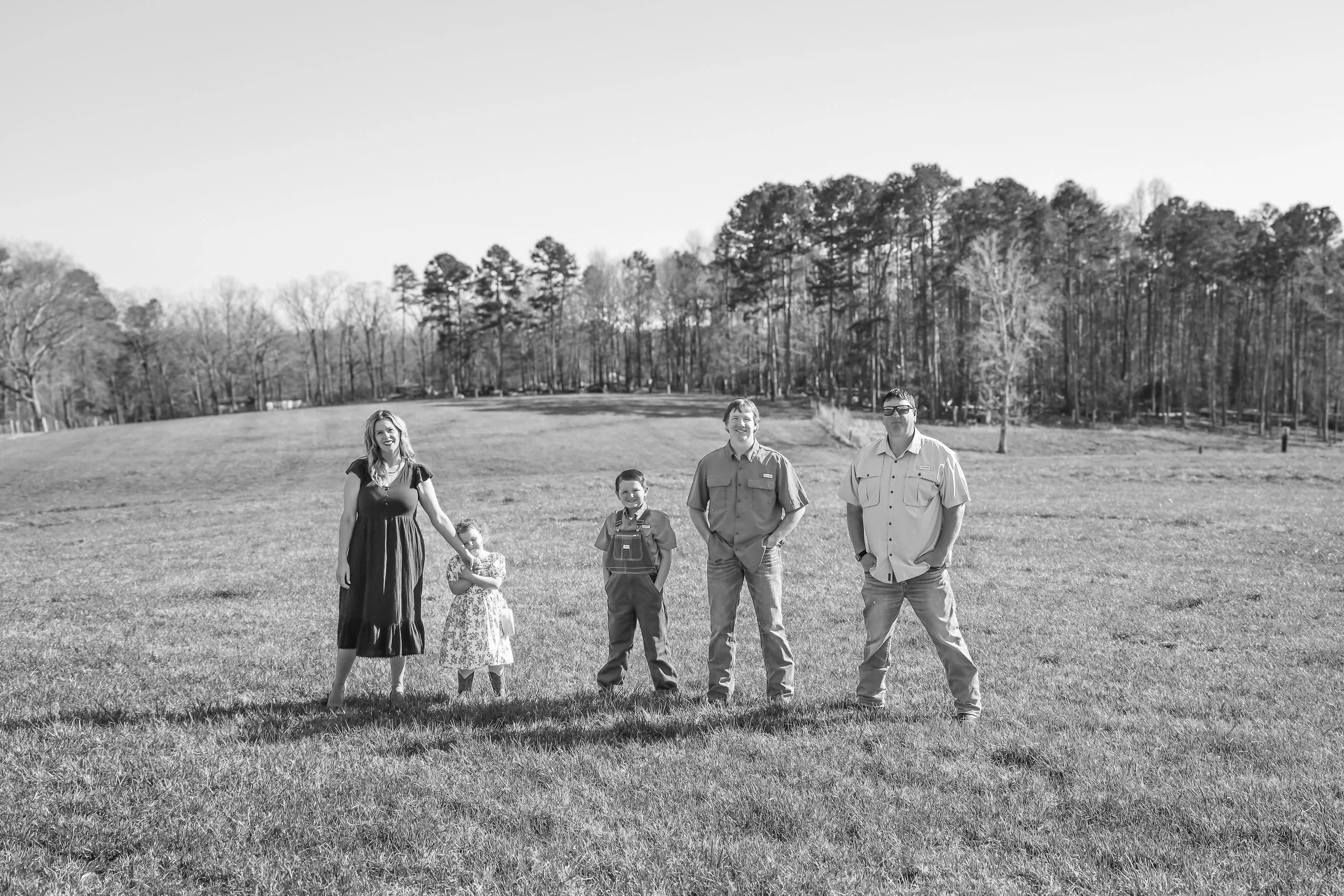 A black and white photo of a family standing on a grassy field with trees in the background. The family includes two adults and three children, with the adults and older children smiling and the younger girl holding hands with the woman.