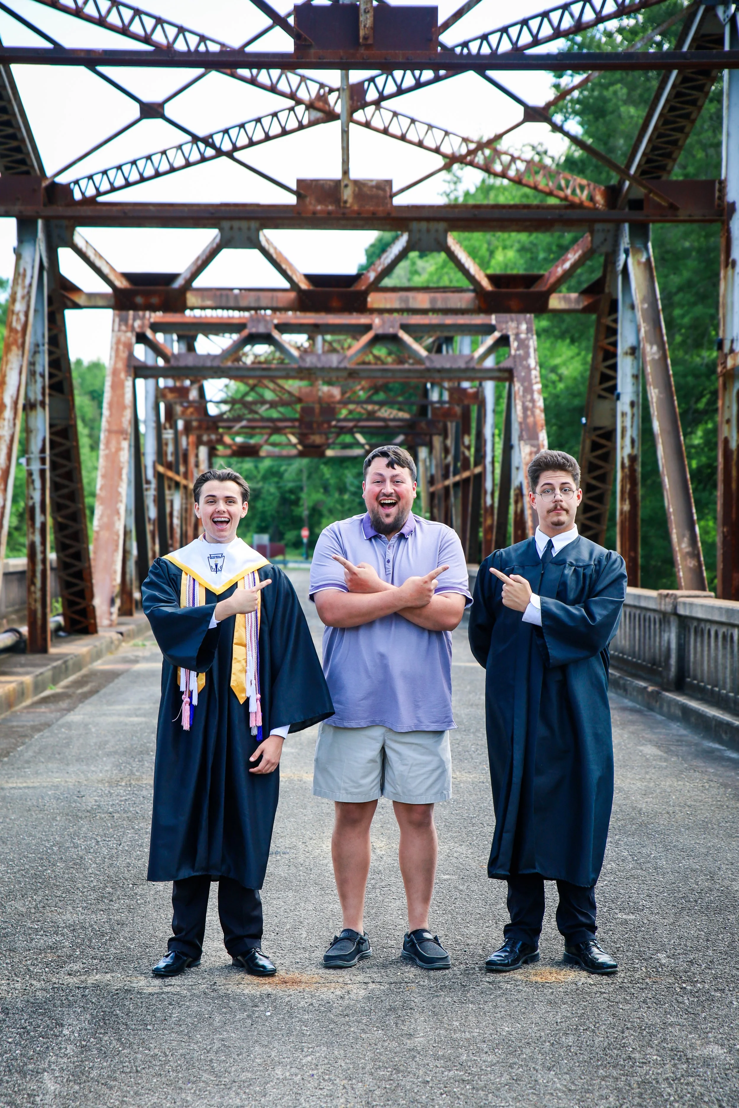 Three young men standing on a bridge, two dressed in graduation gowns and caps, smiling and pointing at the man in the middle who is wearing a casual polo shirt and shorts, under a rusted metal bridge with green trees in the background.