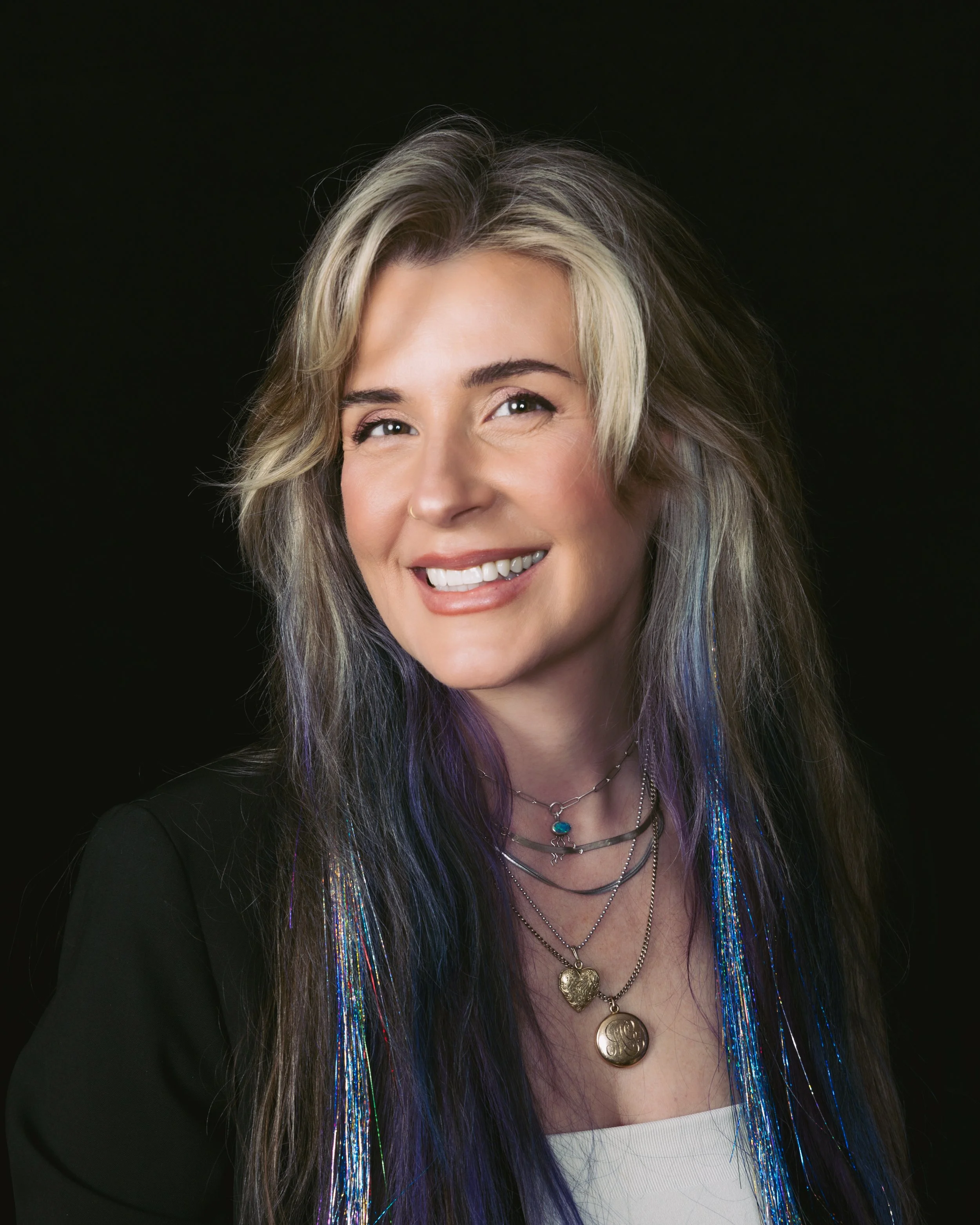 A smiling woman with long, multicolored hair wearing layered necklaces and a black blazer against a black background.
