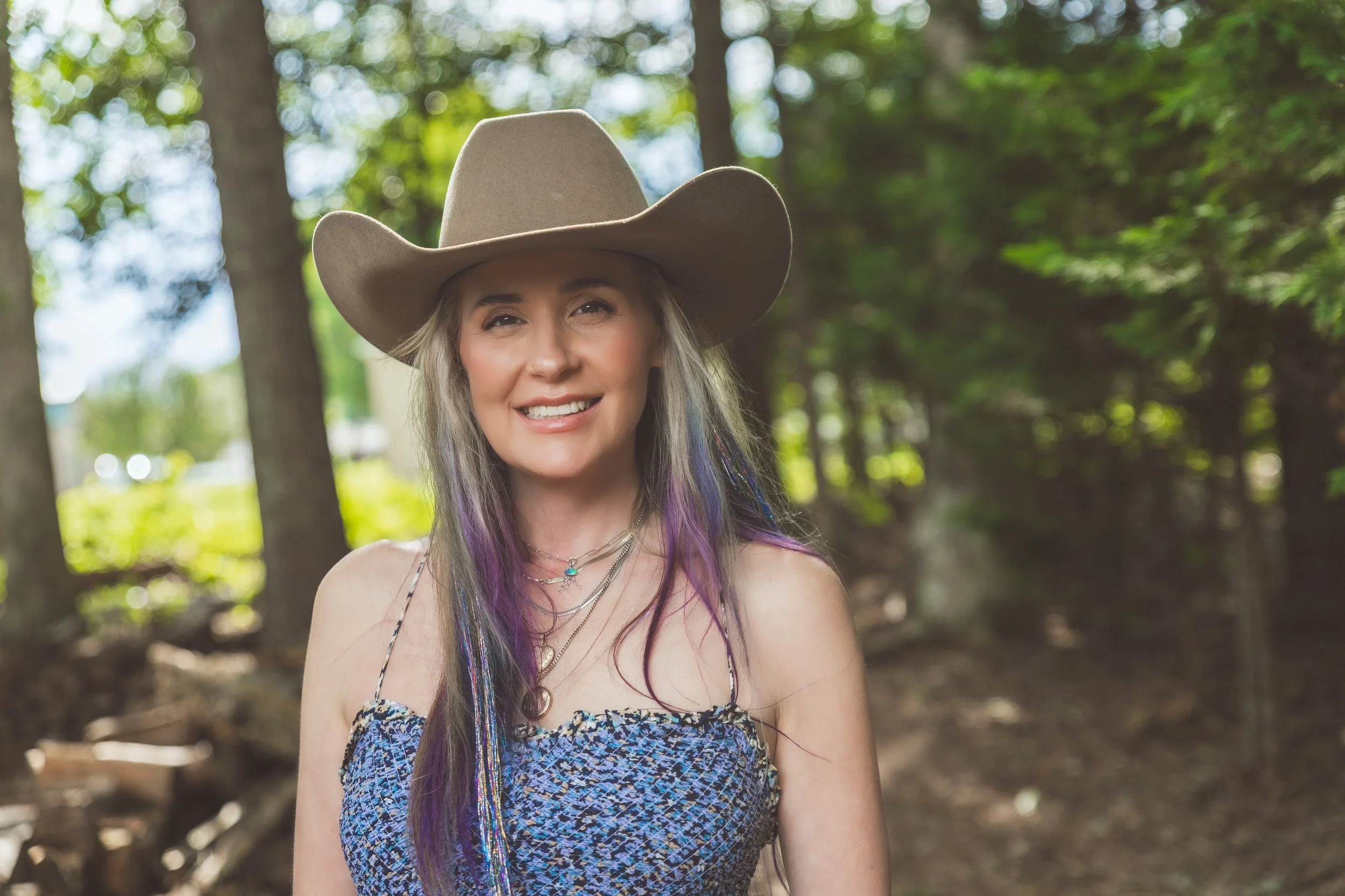 A woman smiling outdoors in a forest setting, wearing a large cowboy hat, patterned dress, and layered necklaces.