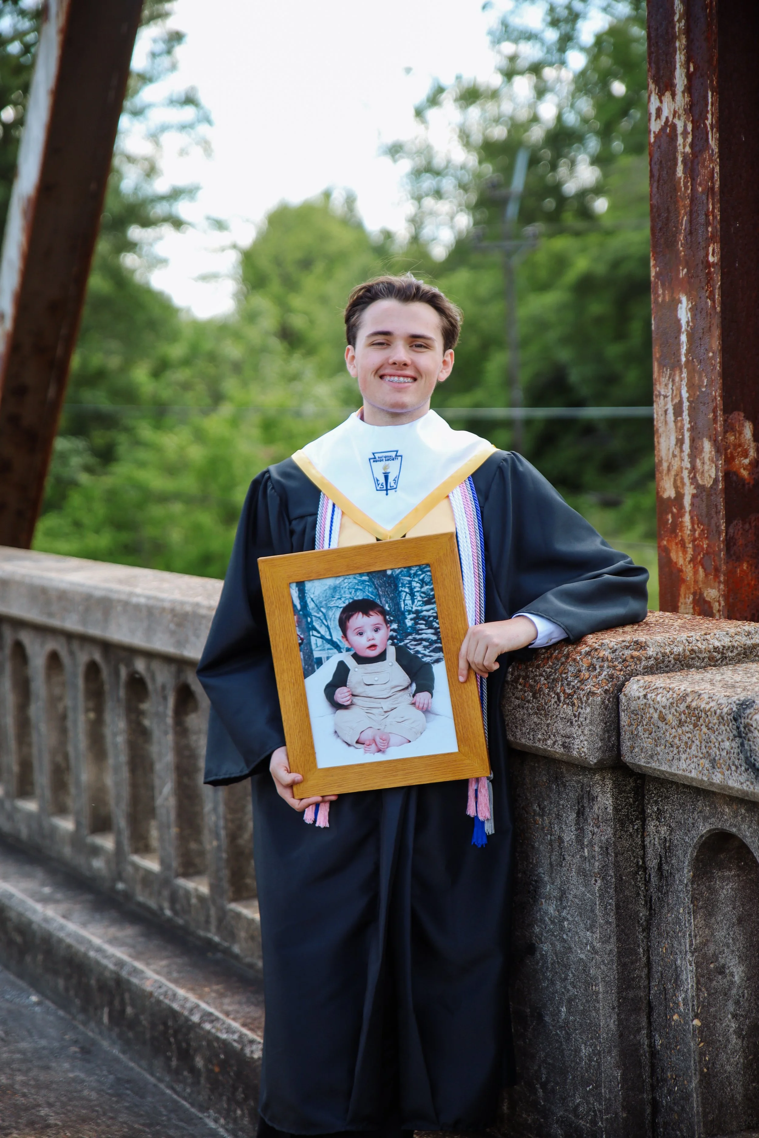 Young man in graduation gown and stole holding a framed photo of a baby boy in outdoor setting.