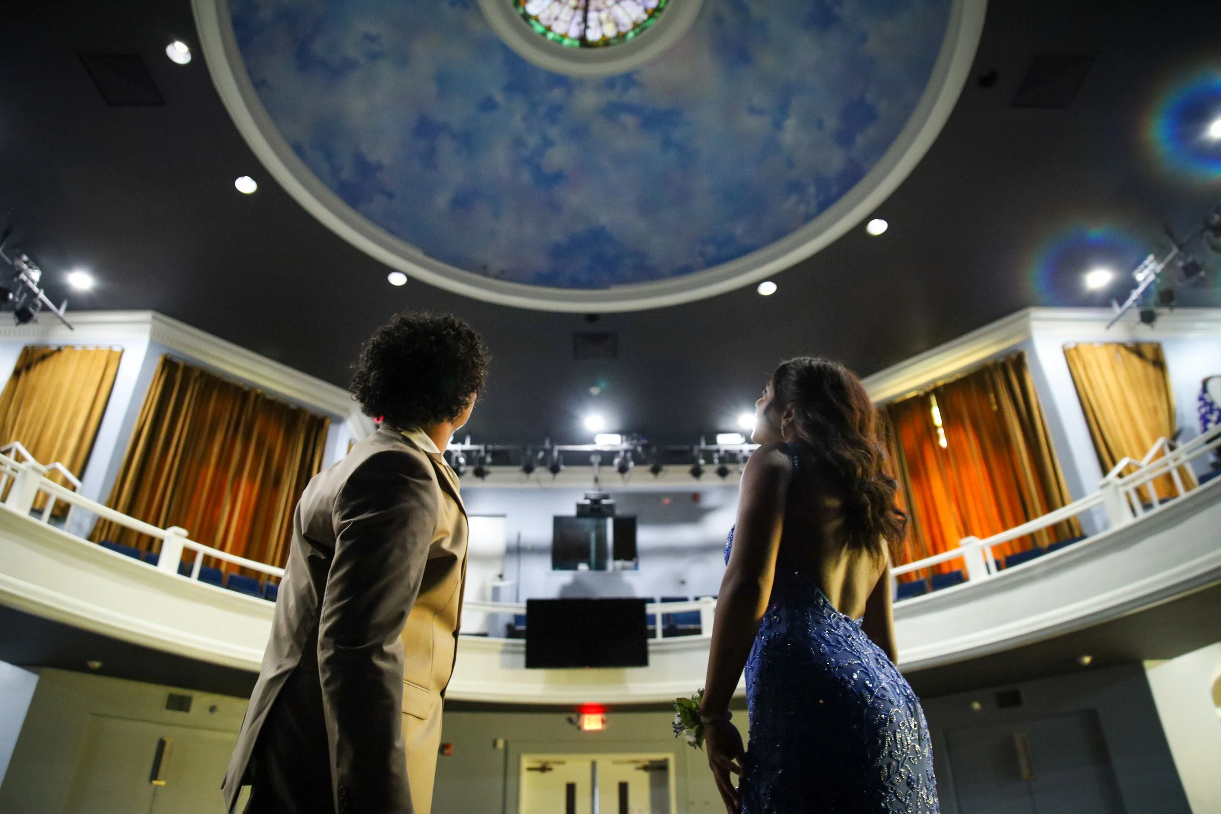 Two people, a man in a tan suit and a woman in a dark blue dress, stand on a stage or dance floor in a ballroom or theater, looking up at the decorated ceiling with clouds.