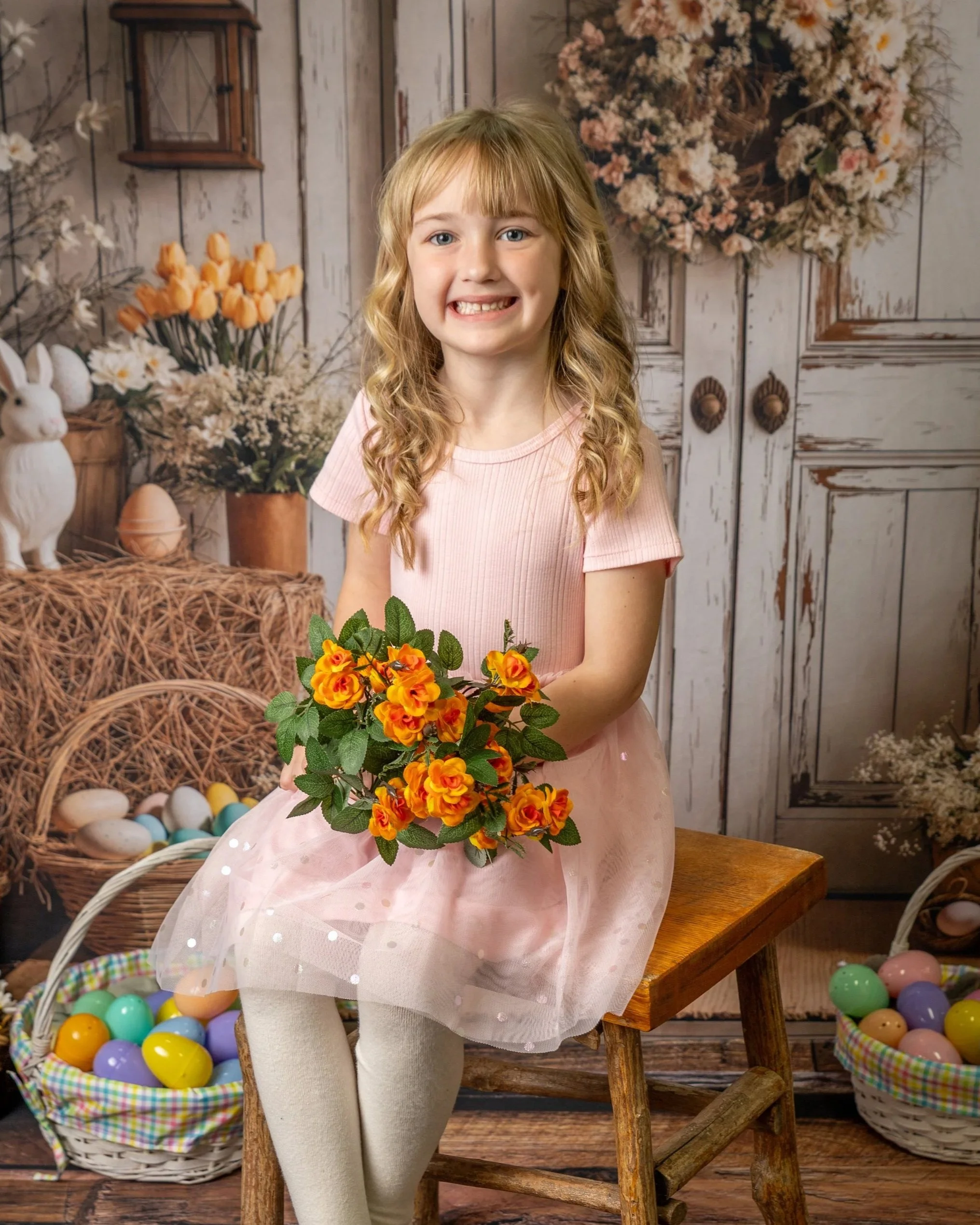 Smiling young boy with short blond hair and light skin, sitting on a wooden stool in front of a fall-themed background with pumpkins and sunflowers, and a rustic white wooden wall.