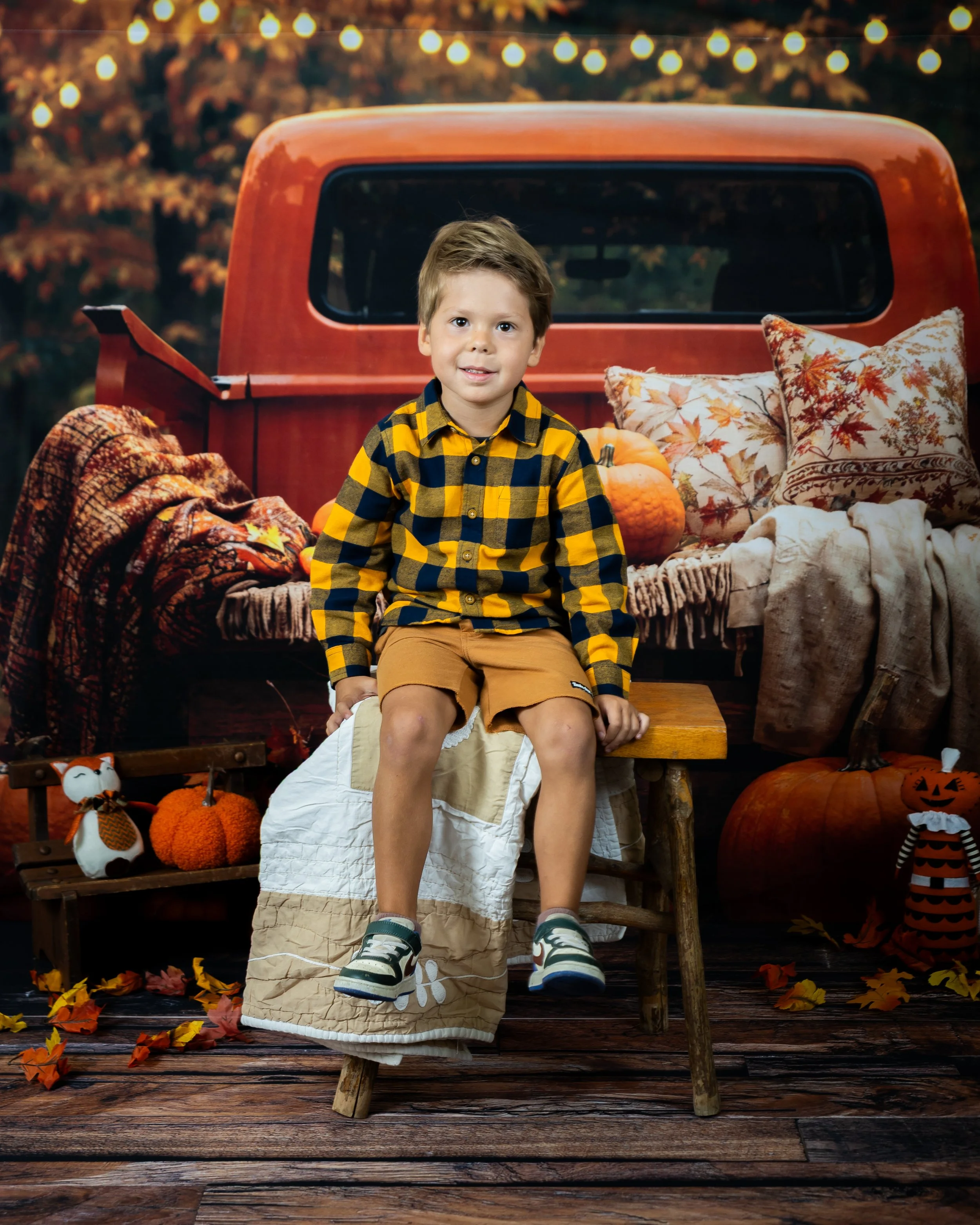 Smiling young boy with short blond hair and light skin, sitting on a wooden stool in front of a fall-themed background with pumpkins and sunflowers, and a rustic white wooden wall.