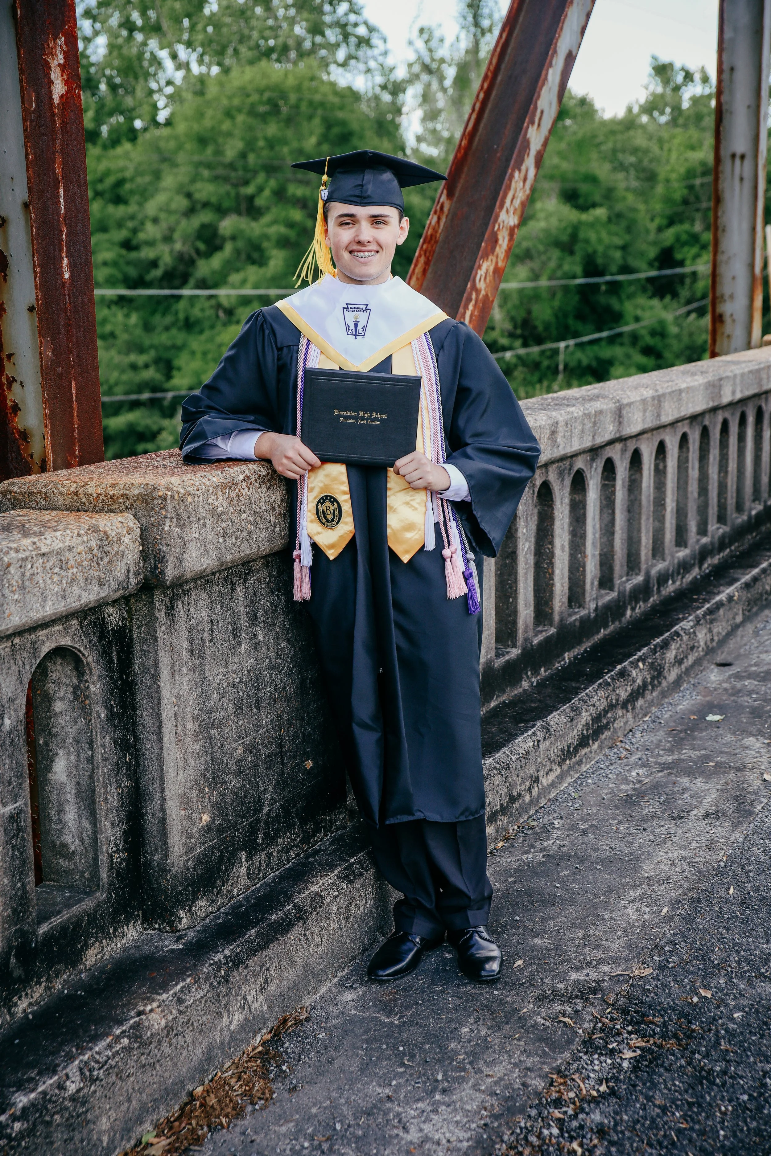 A young man in a graduation gown and cap holding a diploma standing on a bridge with a stone railing and green trees in the background.
