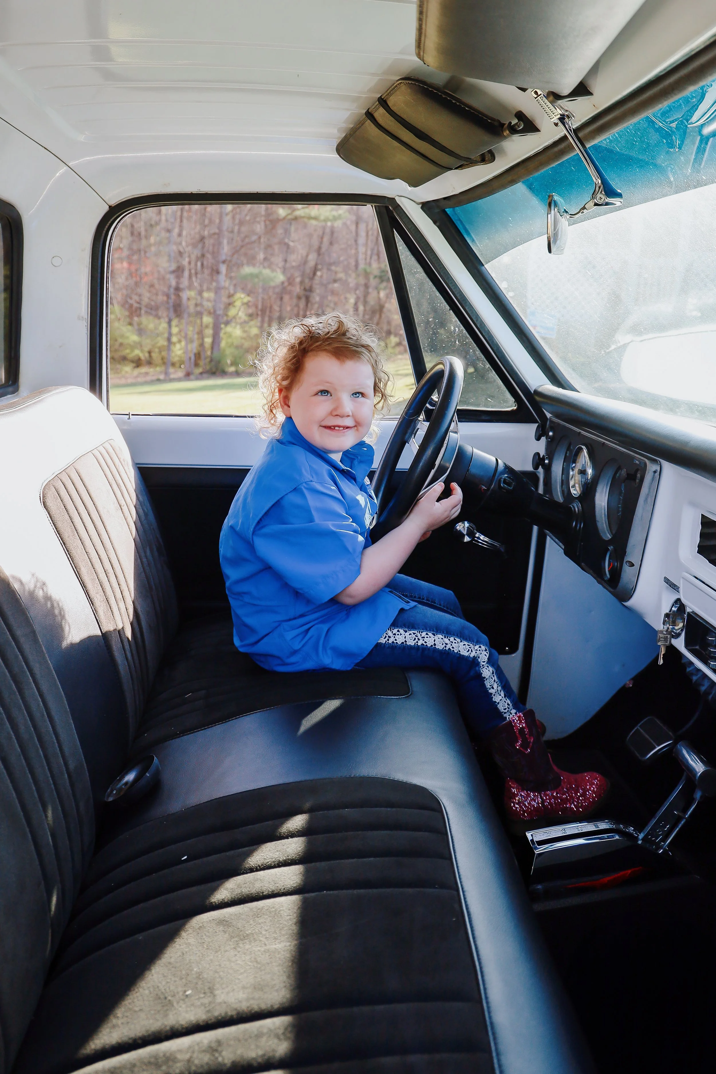 A young girl with curly hair sitting in the driver's seat of a vintage white truck, holding the steering wheel, smiling, wearing a blue shirt, jeans with lace detail, and red glitter boots, with a background of trees and grass seen through the window