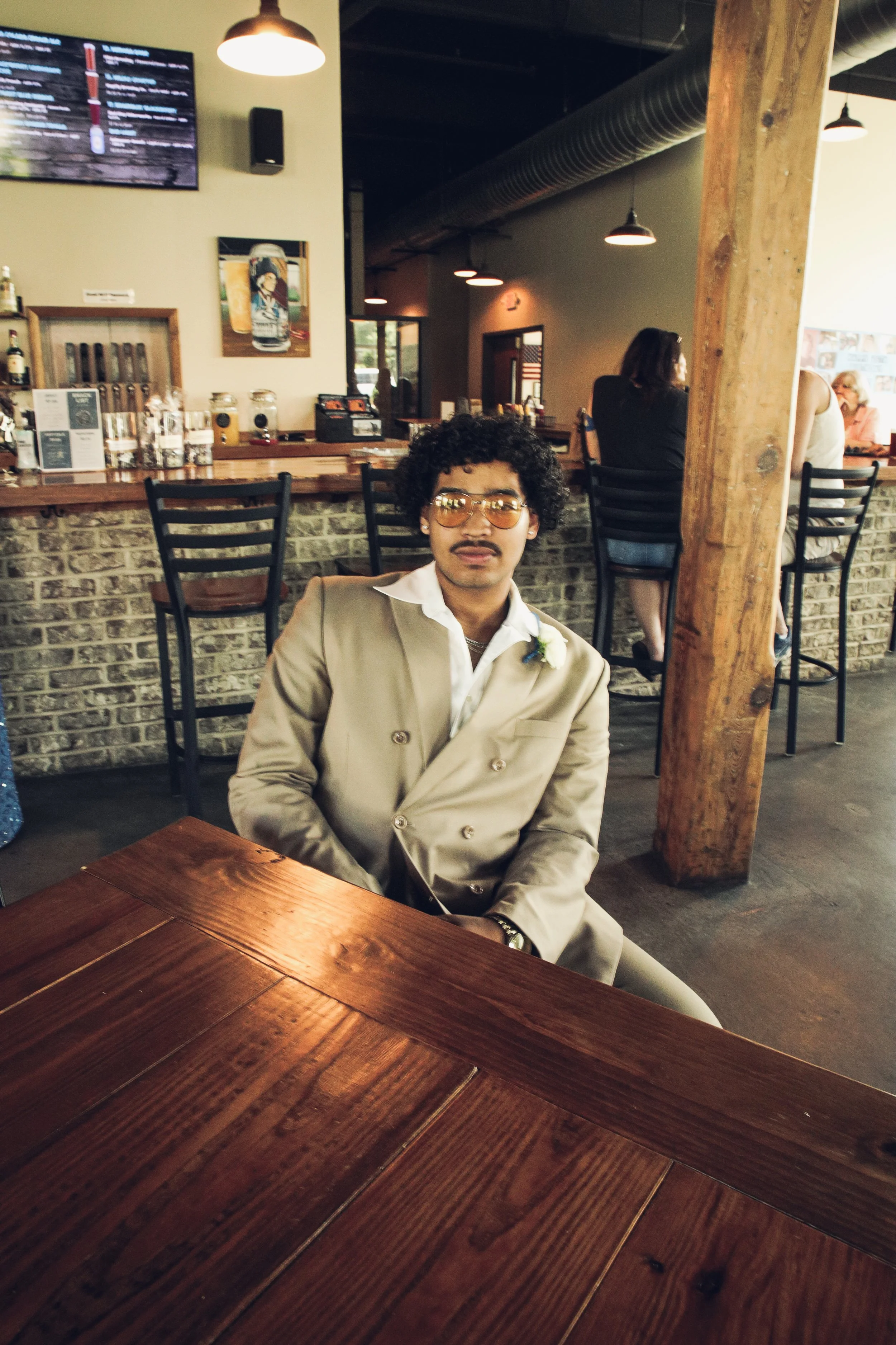 A man with curly black hair and glasses sits at a wooden table in a modern restaurant with exposed brick and wood accents, dressed in a beige double-breasted suit with a white shirt and a small boutonniere.