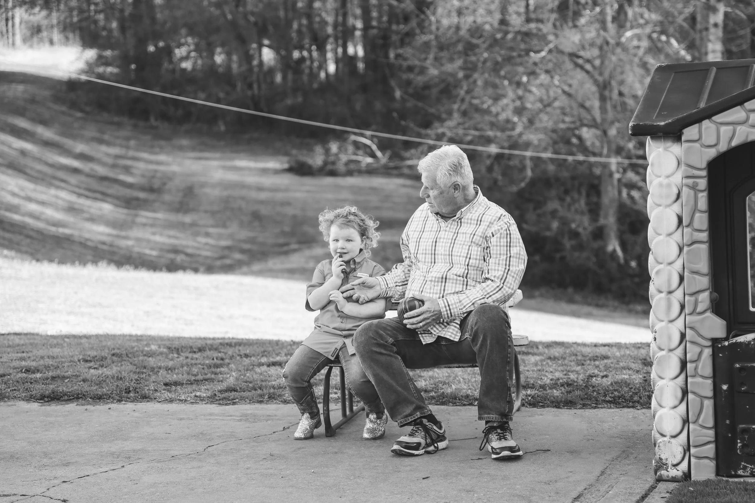 A young child and an older man sitting on a park bench outdoors, with the man holding an object in his hand, and the child holding his hand, beside a small playhouse. The background shows a grassy area and trees.
