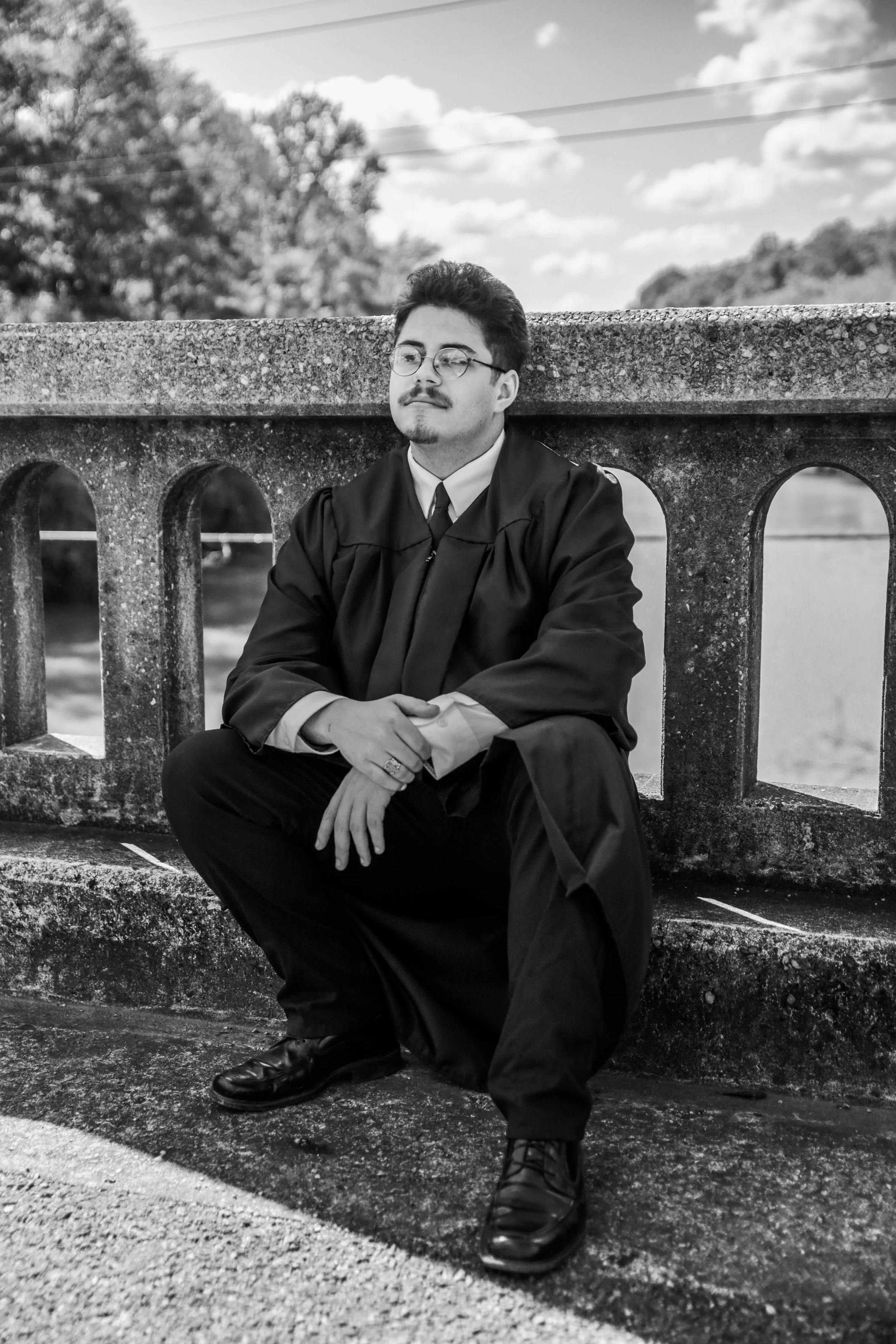 A young man dressed in a graduation gown and cap, sitting on a stone bridge with trees and a cloudy sky in the background.