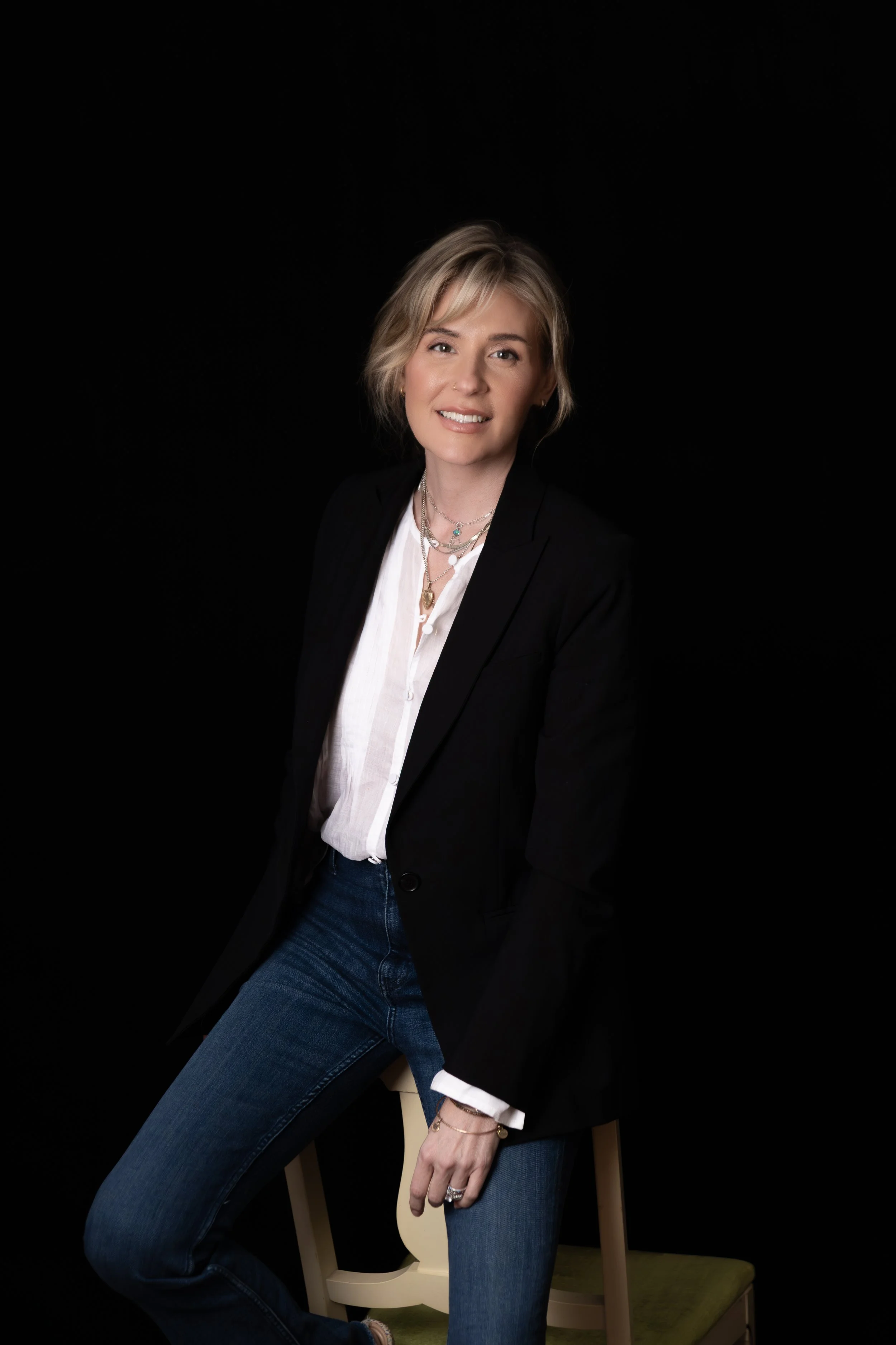 A woman with short blonde hair, wearing a black blazer, white blouse, and blue jeans, sitting on a small wooden chair with a green cushion against a black background.