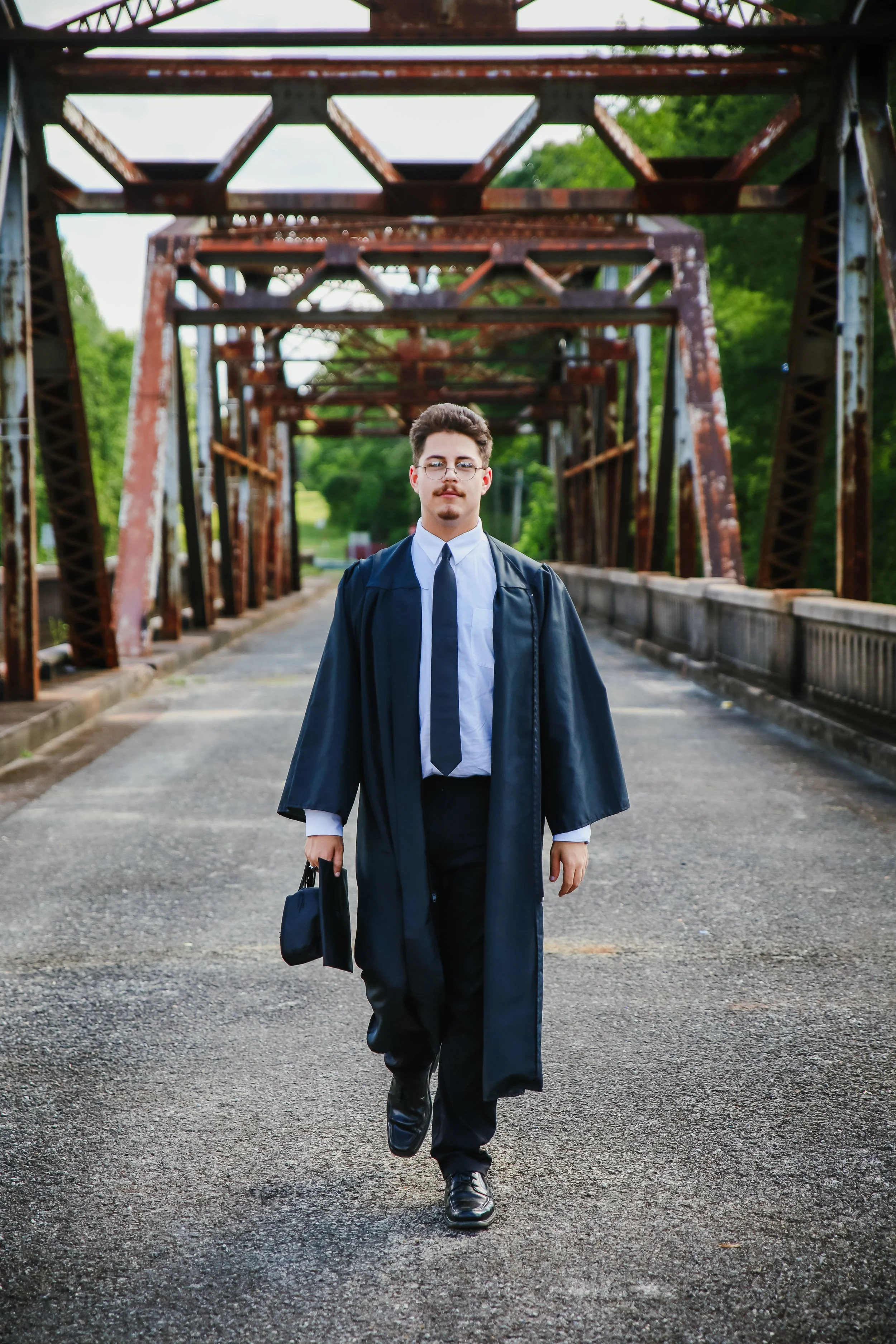 A young man in a cap and gown walking on a bridge with a plate of steel beams overhead.