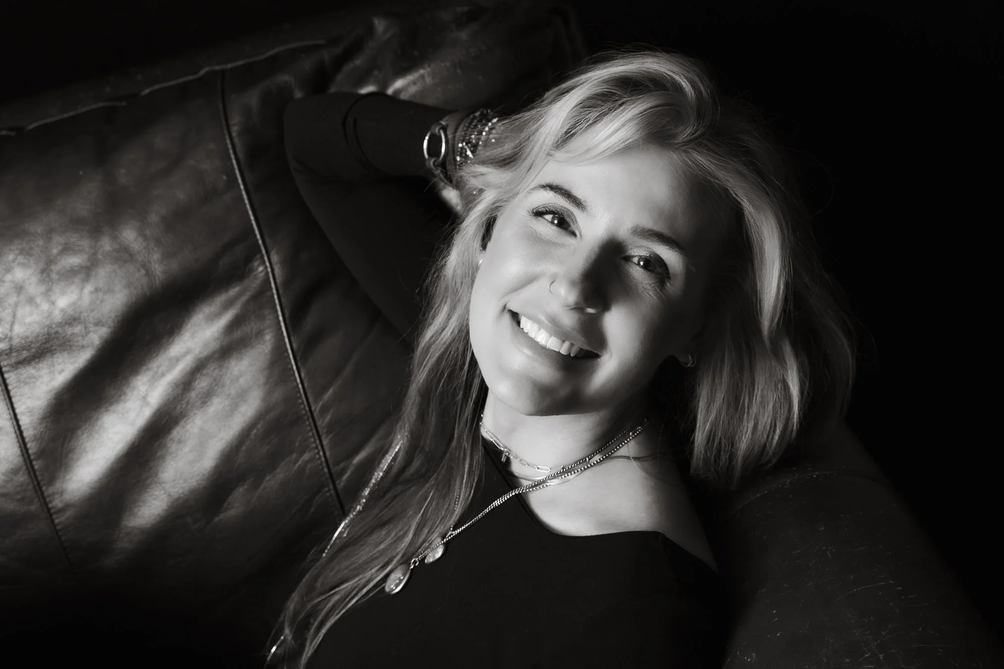 Black and white photo of a woman with wavy hair, smiling and lying on a leather couch, looking up at the camera.