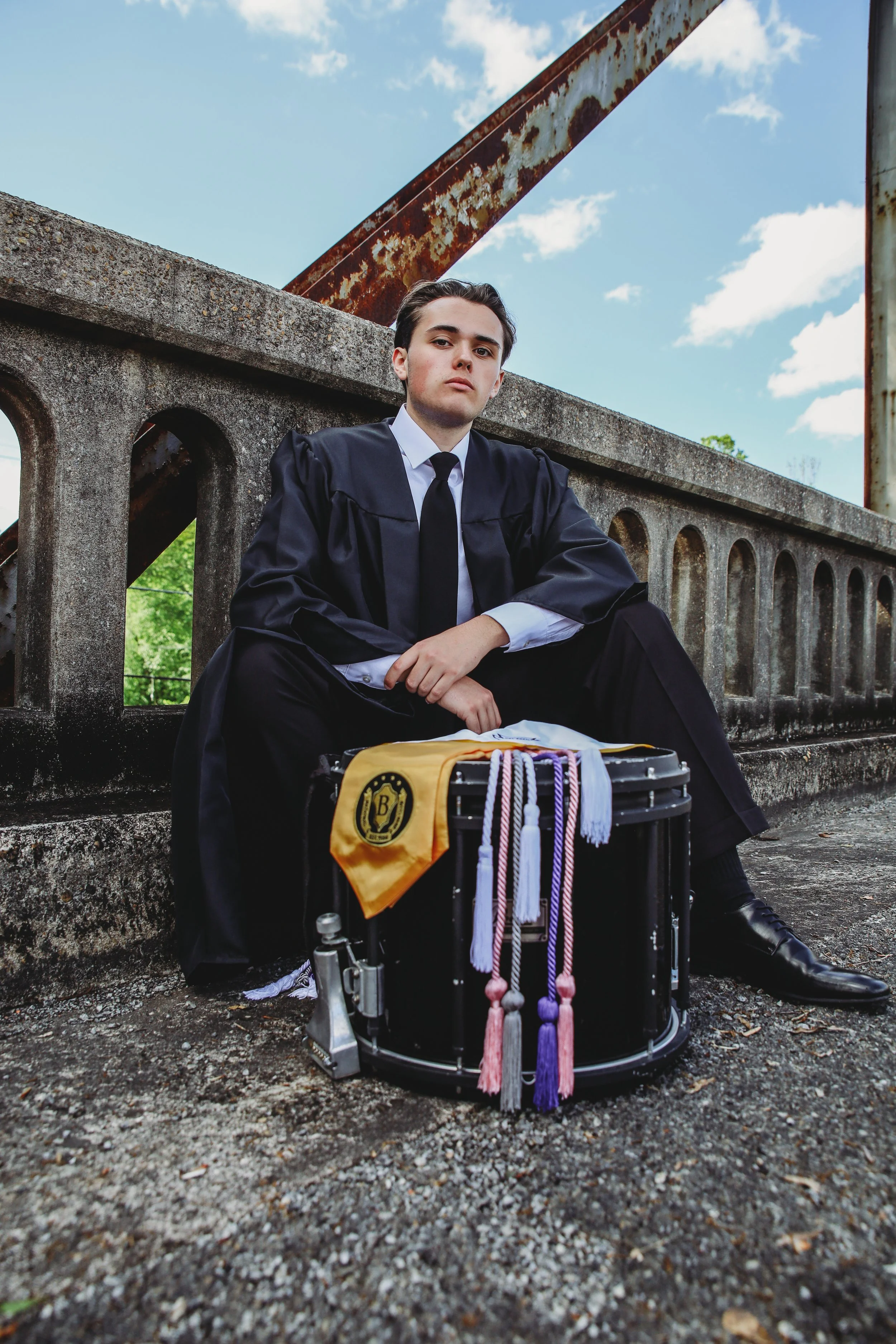 A young man dressed in a graduation cap and gown sitting on a concrete bridge railing with a drum, decorated with tassels and honor cords, in front of him. The sky is blue with clouds and trees are visible in the background.