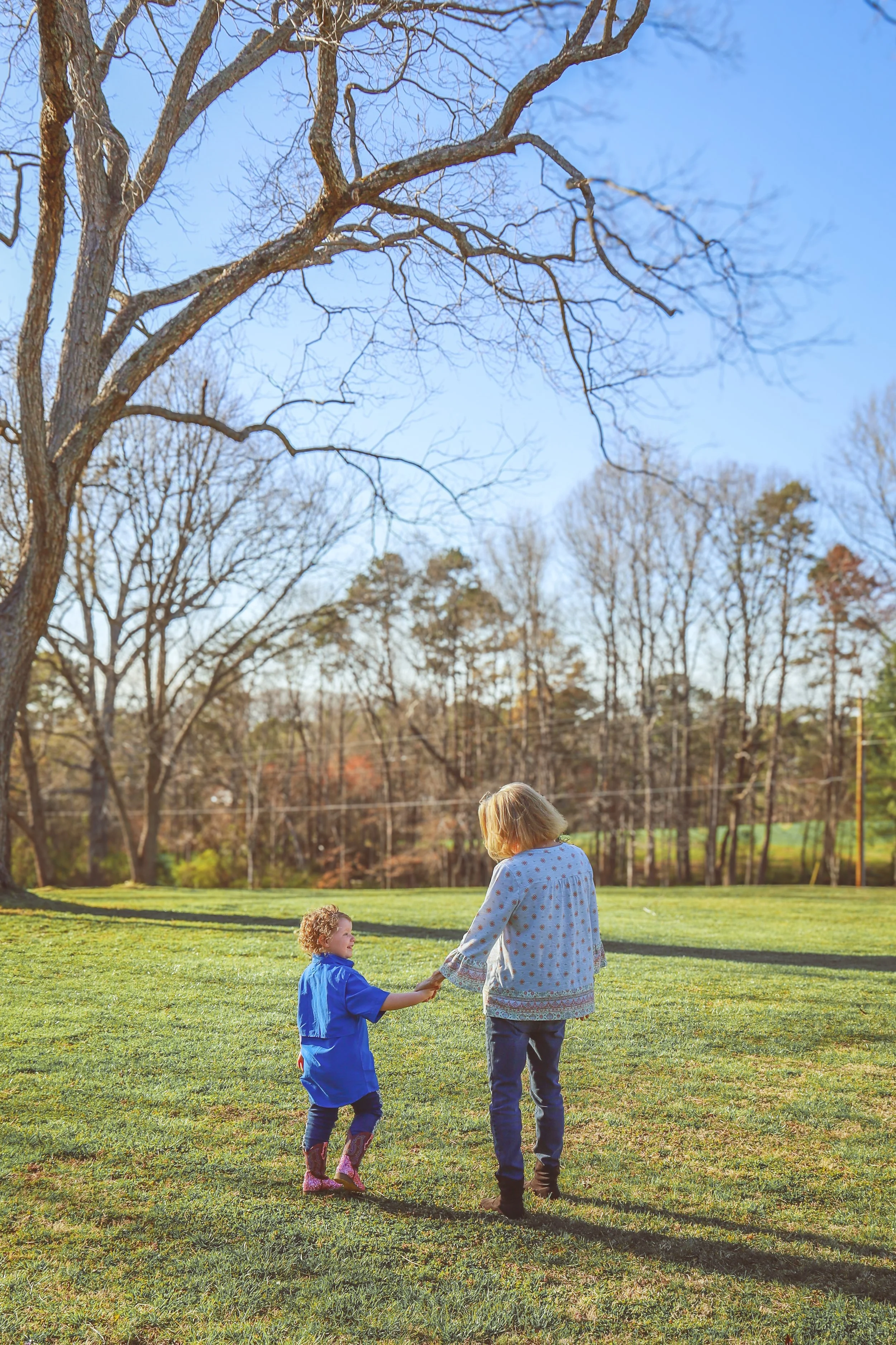 An older woman and a young girl holding hands while walking on a grassy field with trees in the background on a sunny day.