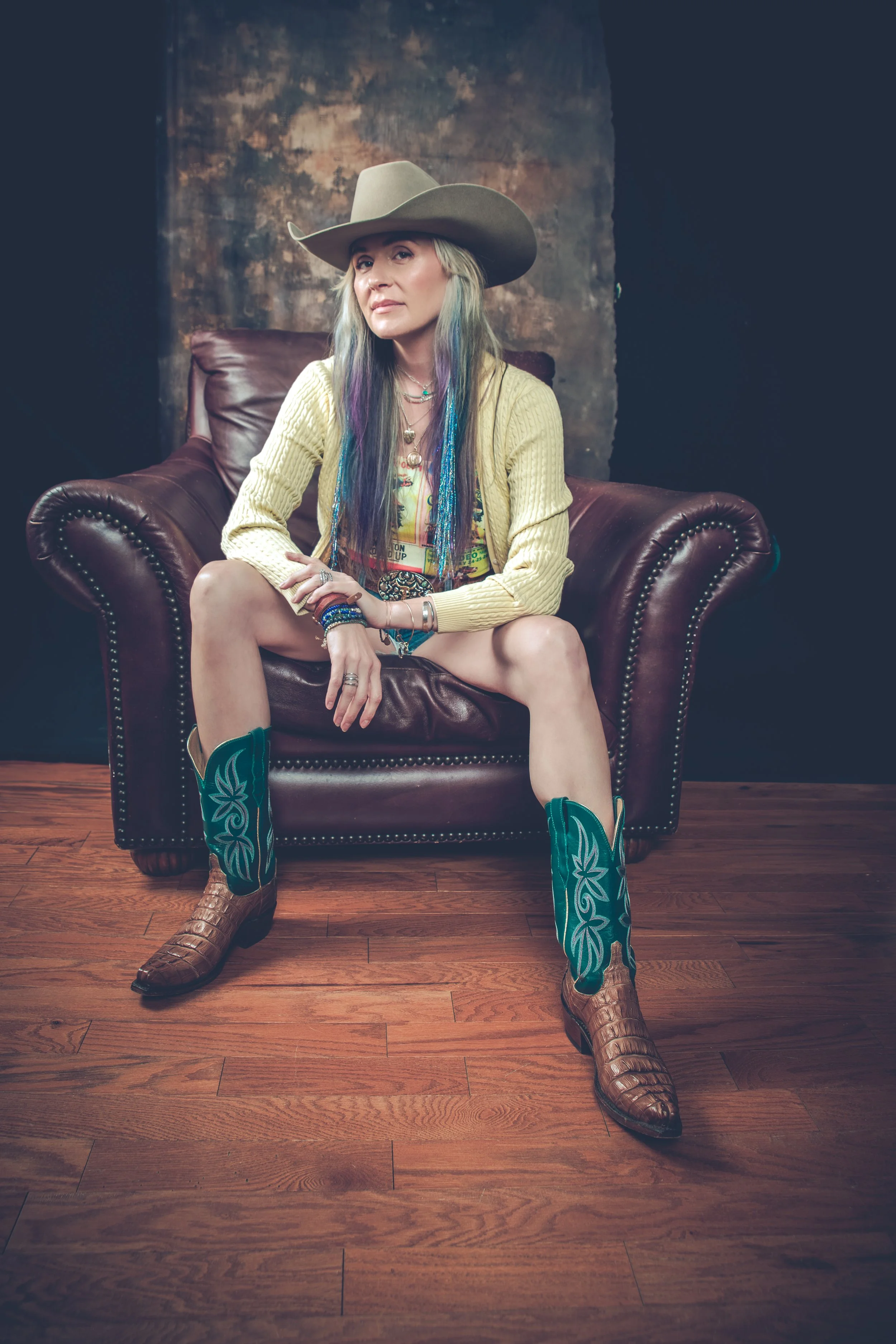 A woman with colorful hair and multiple necklaces sits on a leather armchair, wearing a cowboy hat, yellow cardigan, printed shirt, cowboy boots with green accents, and denim shorts, in a room with a wooden floor and a textured wall background.