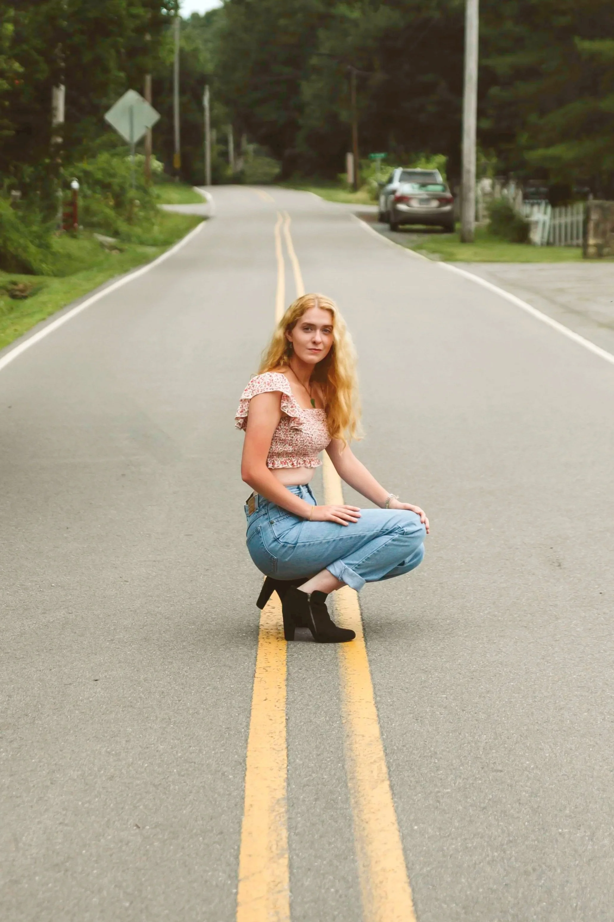 A young woman with long, wavy blonde hair, wearing a cropped floral top, light blue jeans, and black heeled ankle boots, standing in the middle of a two-lane residential street surrounded by greenery and trees.