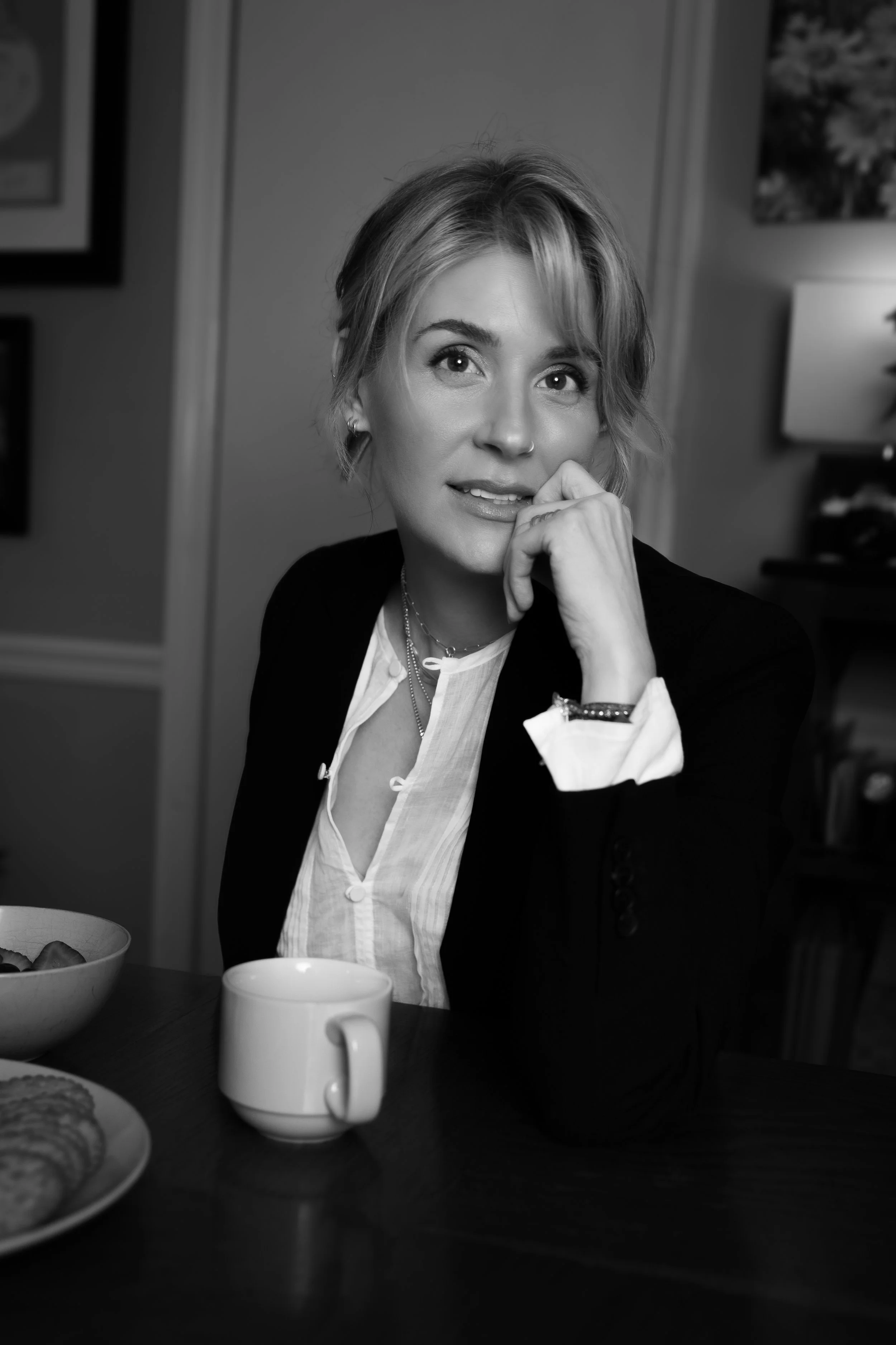 Black and white photo of a woman seated at a table, with a coffee mug and a bowl of cookies or snacks in front of her. She has short, wavy hair, and is wearing a dark blazer over a light blouse. She has her hand near her face, with her chin resting o