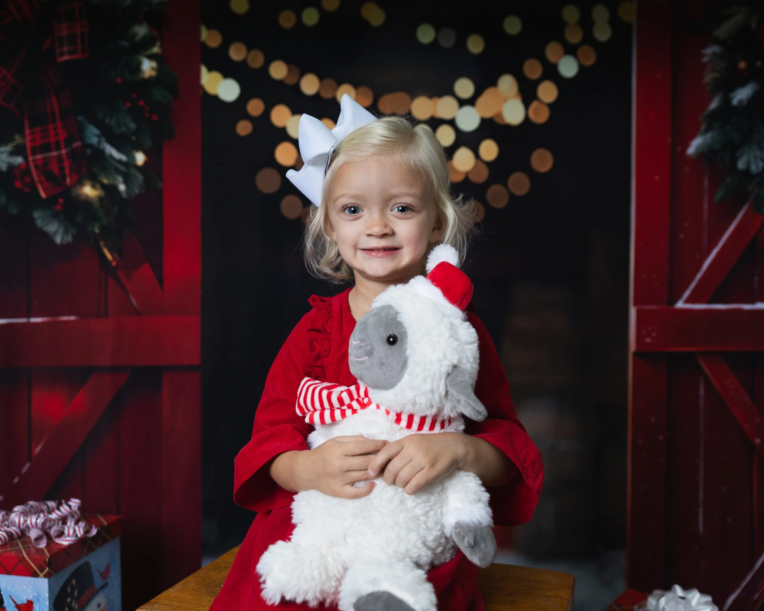 Smiling young boy with short blond hair and light skin, sitting on a wooden stool in front of a fall-themed background with pumpkins and sunflowers, and a rustic white wooden wall.