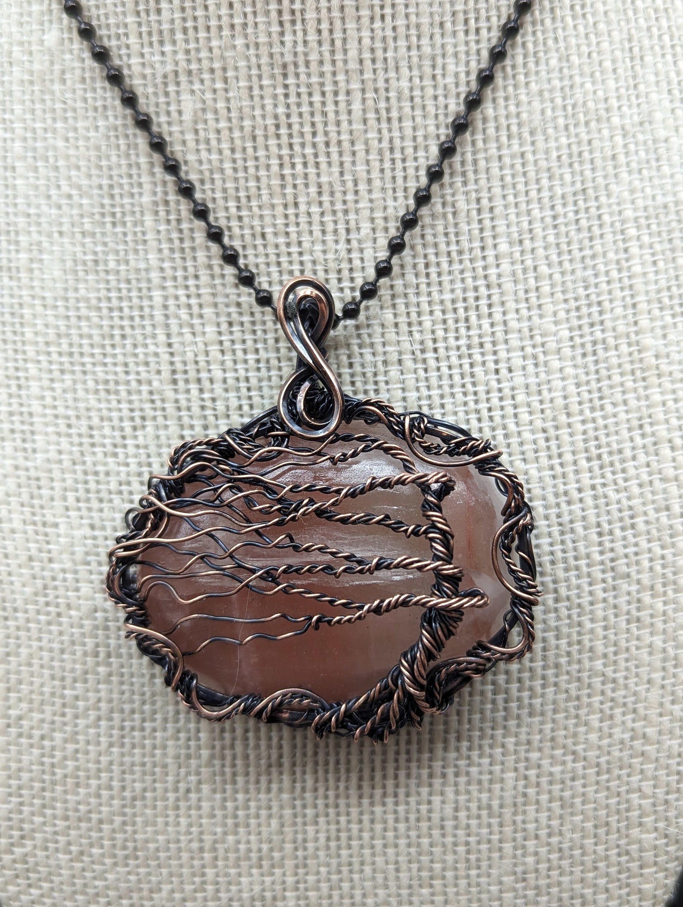 A peach calcite thumb stone copper wire-wrapped pendant featuring a wire wind-swept Tree of Life on the front, surrounded by an elegant wavy twisted wire border and an infinity bail.