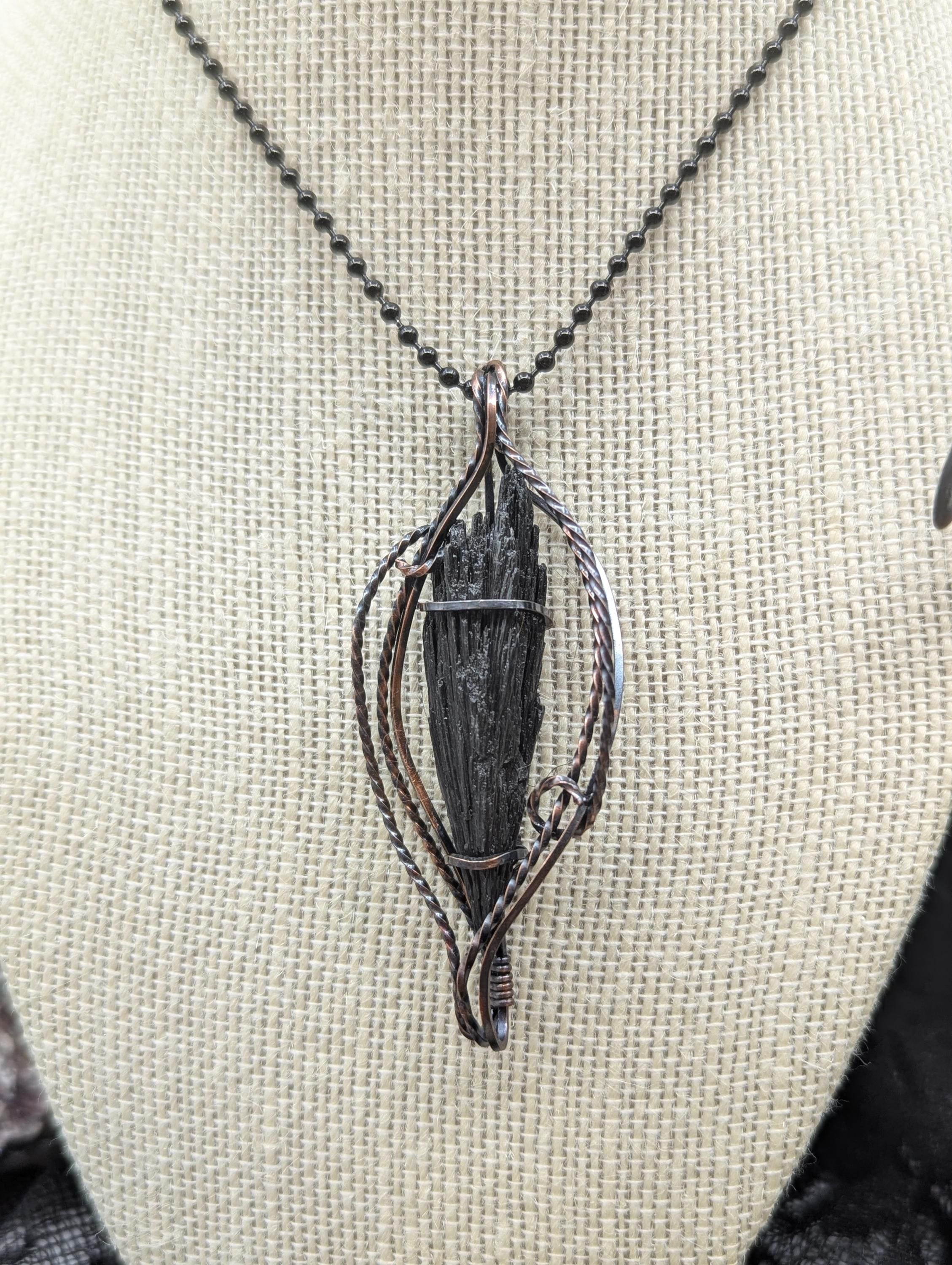 Explore this exquisite Black Kyanite pendant, artfully wrapped in copper wire. Its stunning feathered design and twisted accents elegantly showcase the kyanite as the centerpiece.