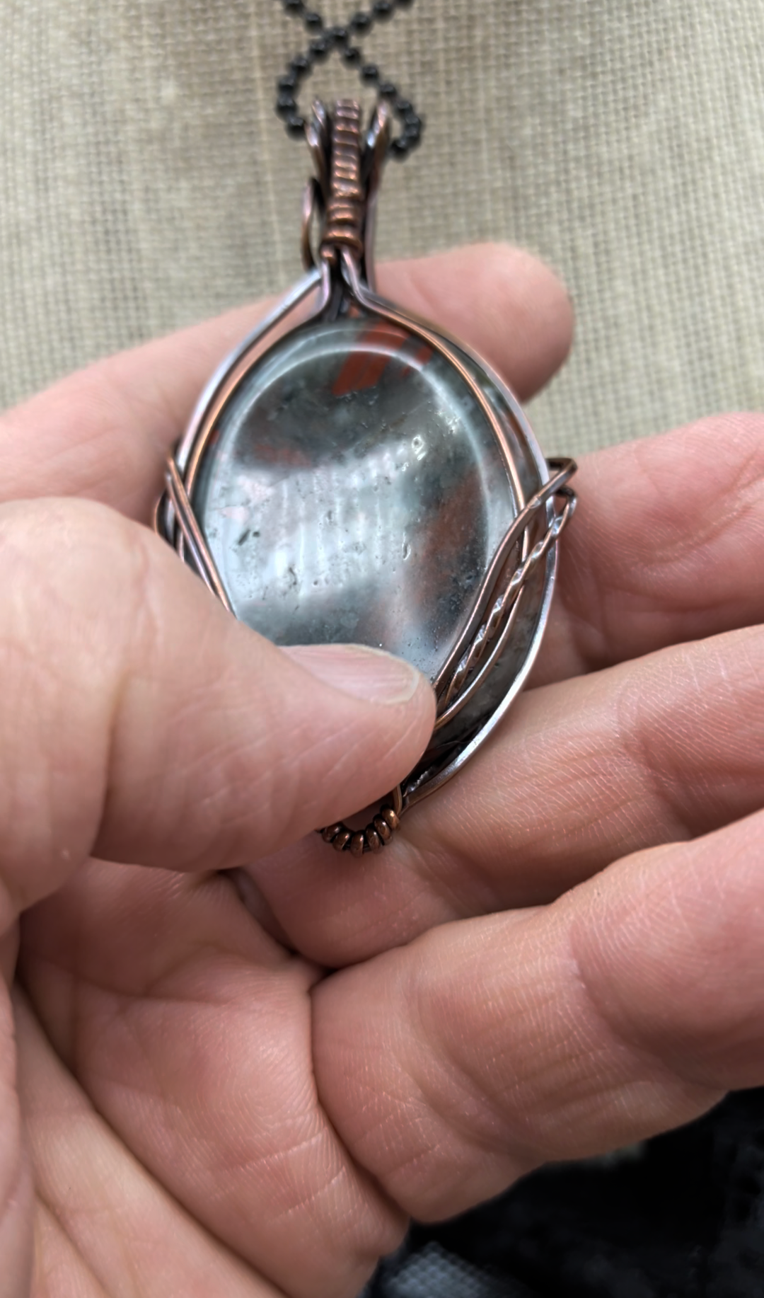 An African Bloodstone Setonite thumb stone copper wire-wrapped pendant featuring a wire swept pattern on the front, surrounded by an elegant wavy twisted wire border.