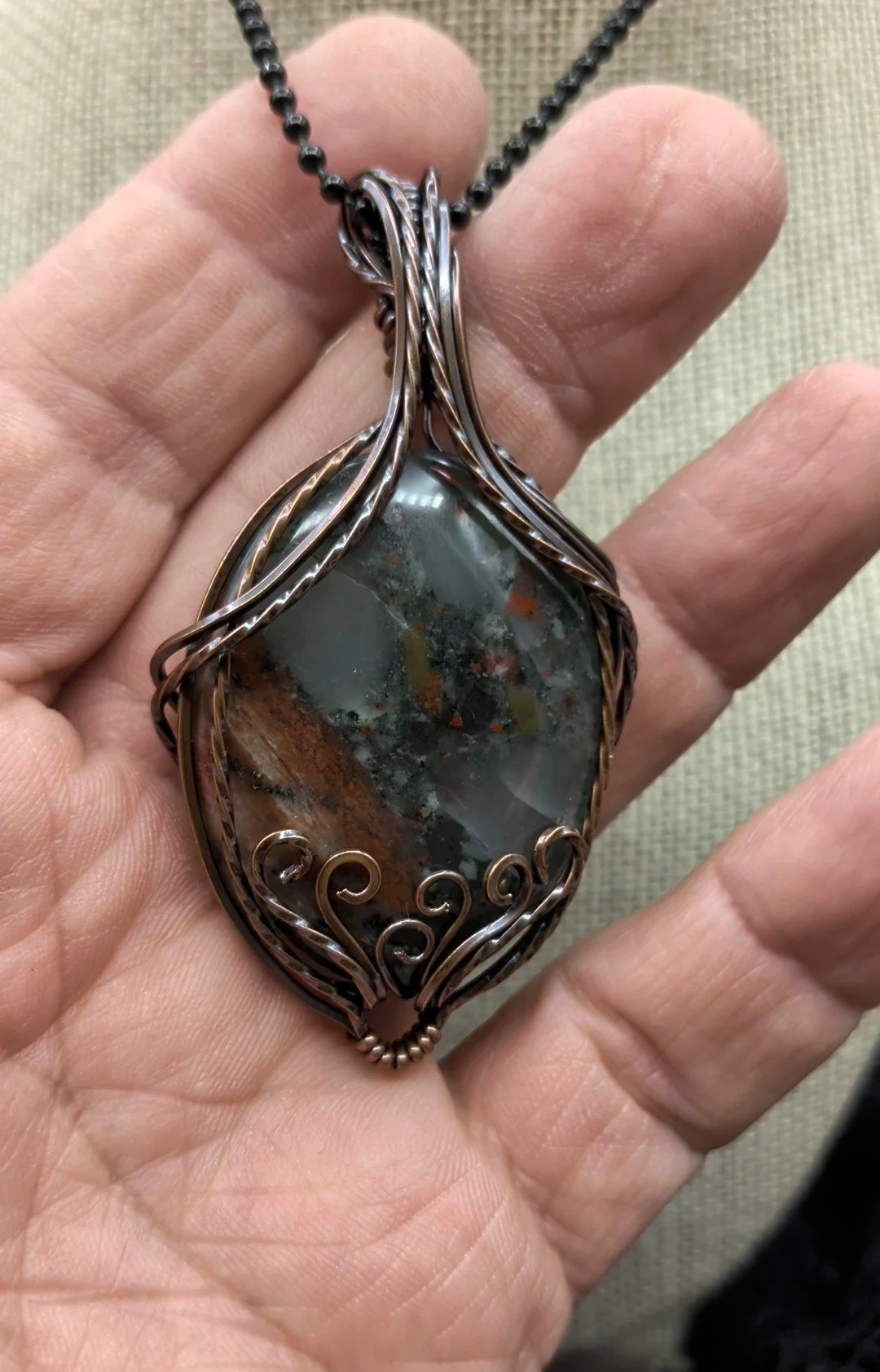 An African Bloodstone Setonite thumb stone copper wire-wrapped pendant featuring a wire swept pattern on the front, surrounded by an elegant wavy twisted wire border.