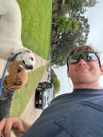 Man taking a selfie with two dogs in a suburban neighborhood park, green grass, trees, parked cars, overcast sky.