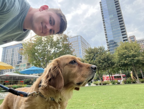 A young man taking a selfie with a golden retriever in a city park, with tall buildings and trees in the background.