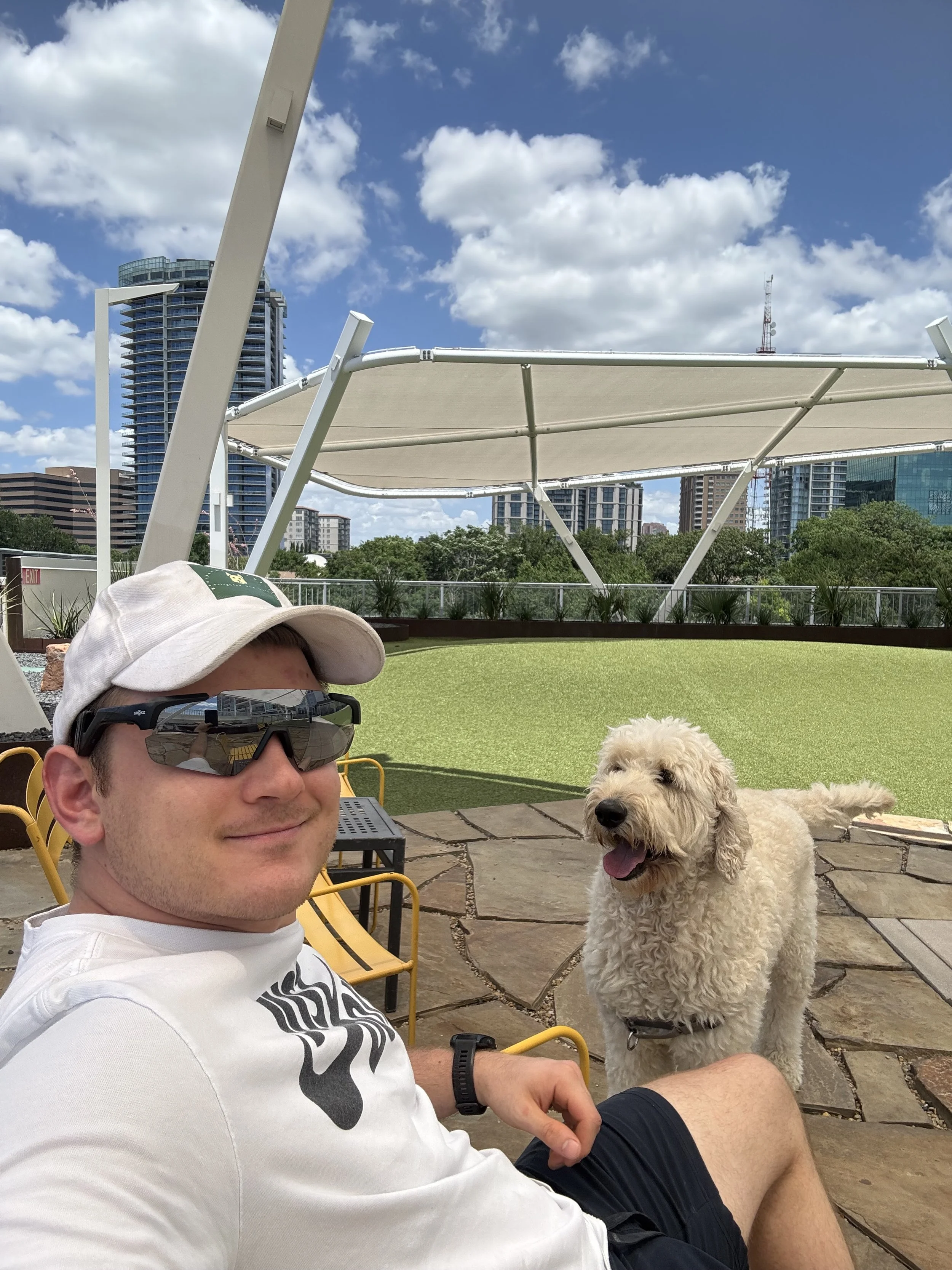 A man wearing a white baseball cap, sunglasses, and a white t-shirt taking a selfie with a happy light-colored dog, possibly a Labradoodle, in an outdoor urban park with a city skyline and a partly cloudy sky in the background.