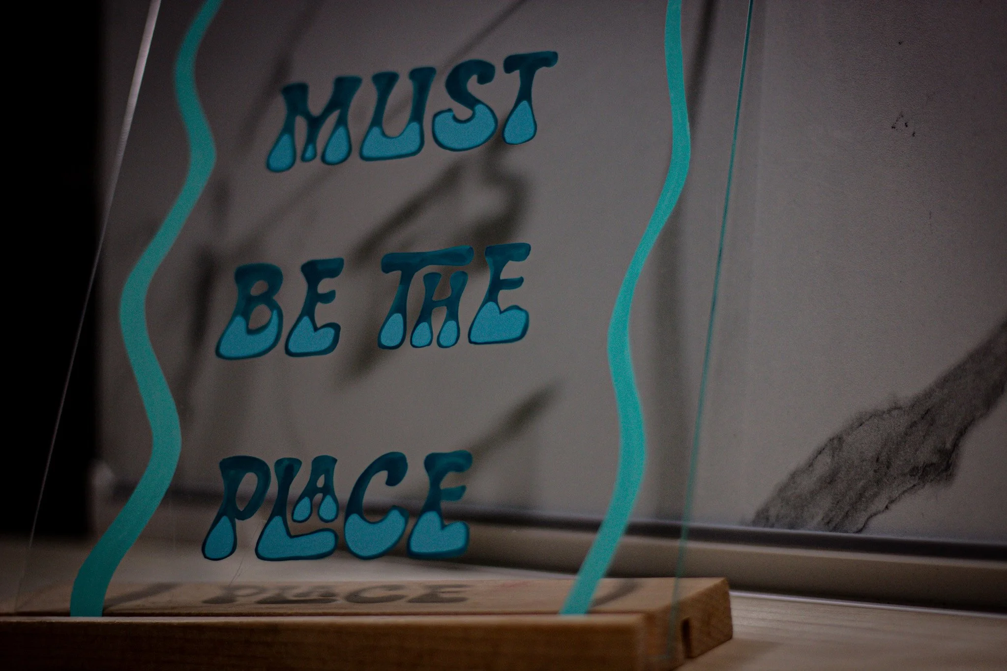 Close-up of a glass sign with the text 'Must Be The Place' written in two tone blue letters, viewed from below.